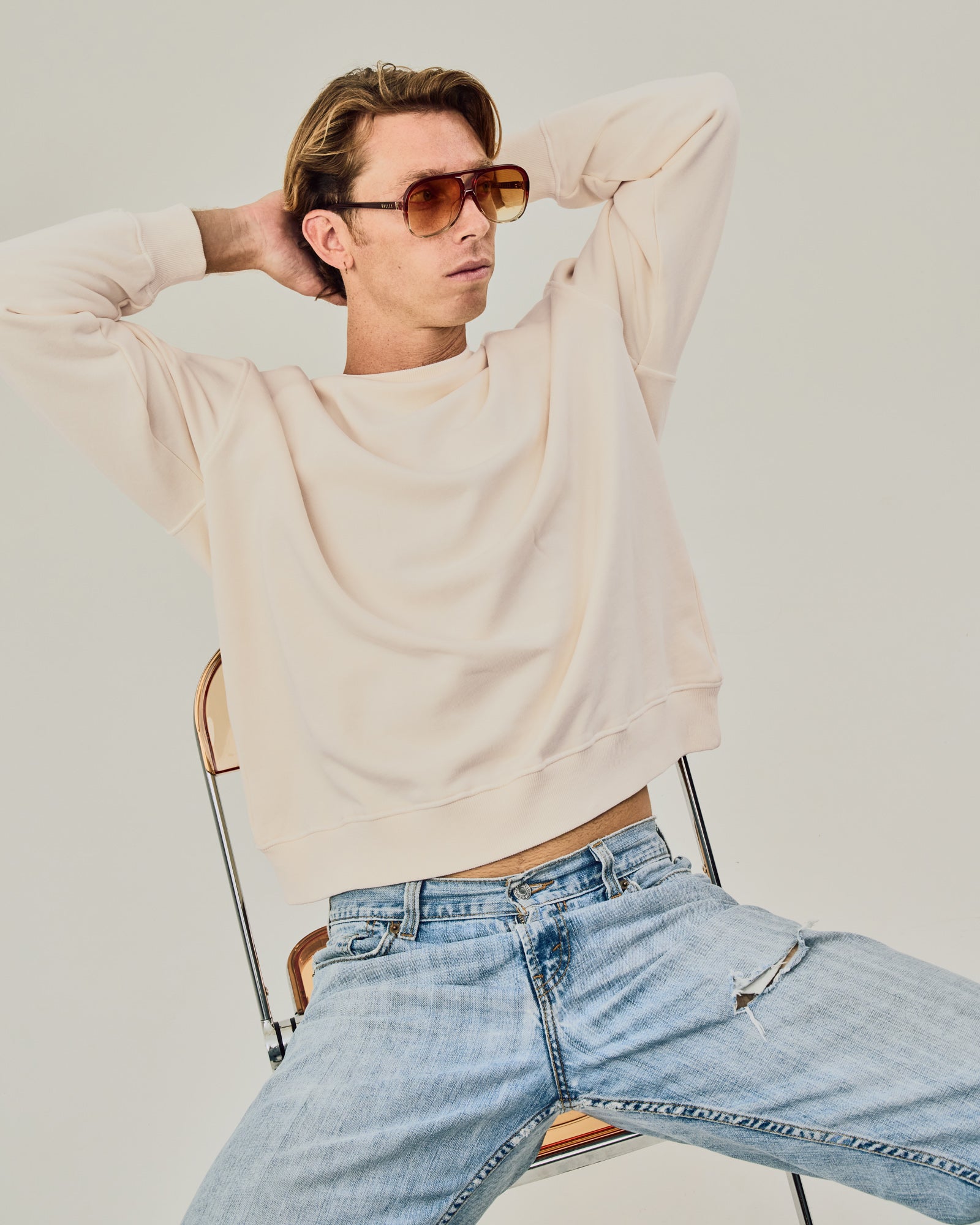 Person wearing a beige sweater and light blue jeans sitting on a chair against a plain background