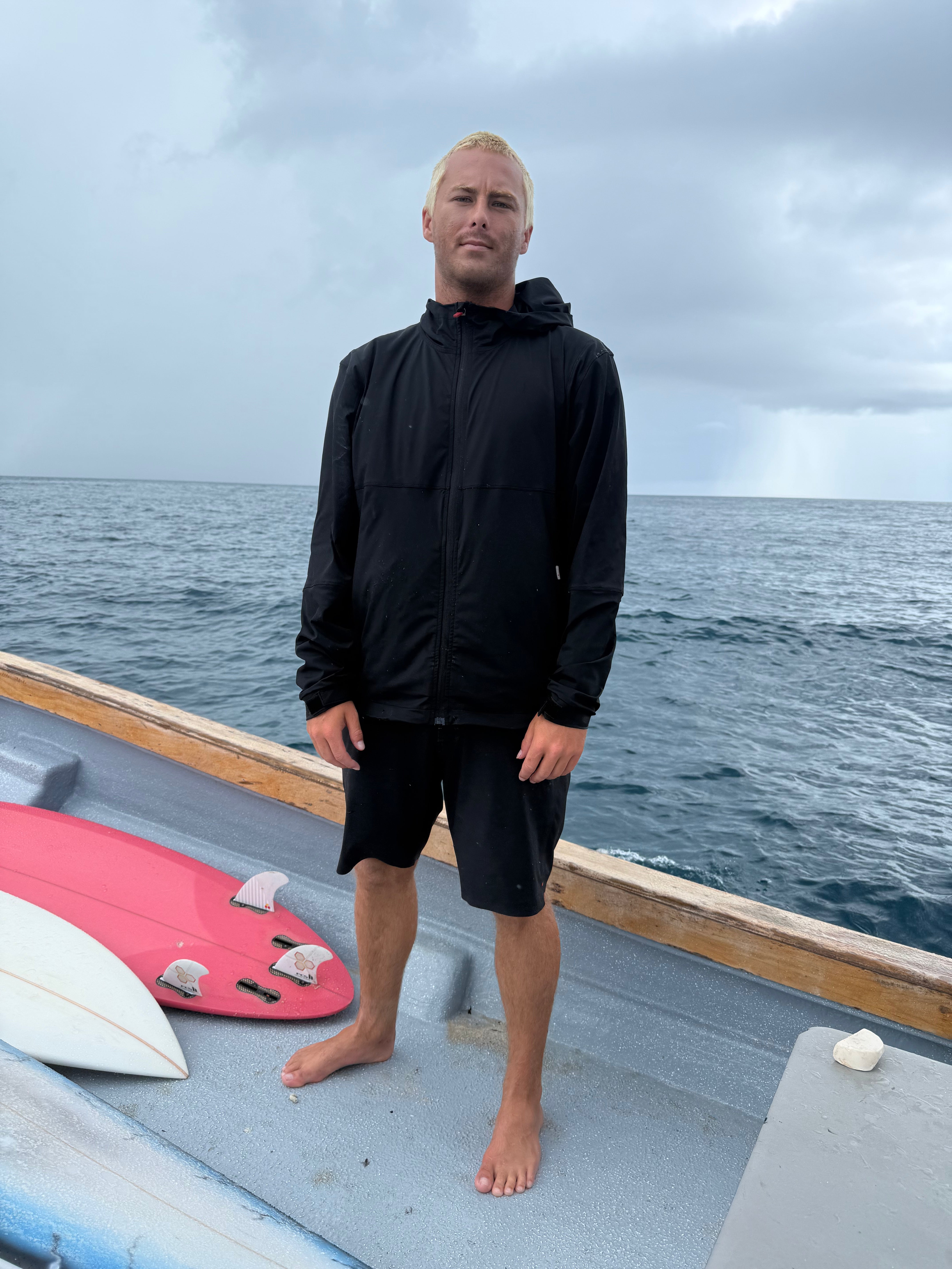 Man standing on a boat with surfboards, wearing a black jacket and shorts, against a cloudy sky and ocean background.