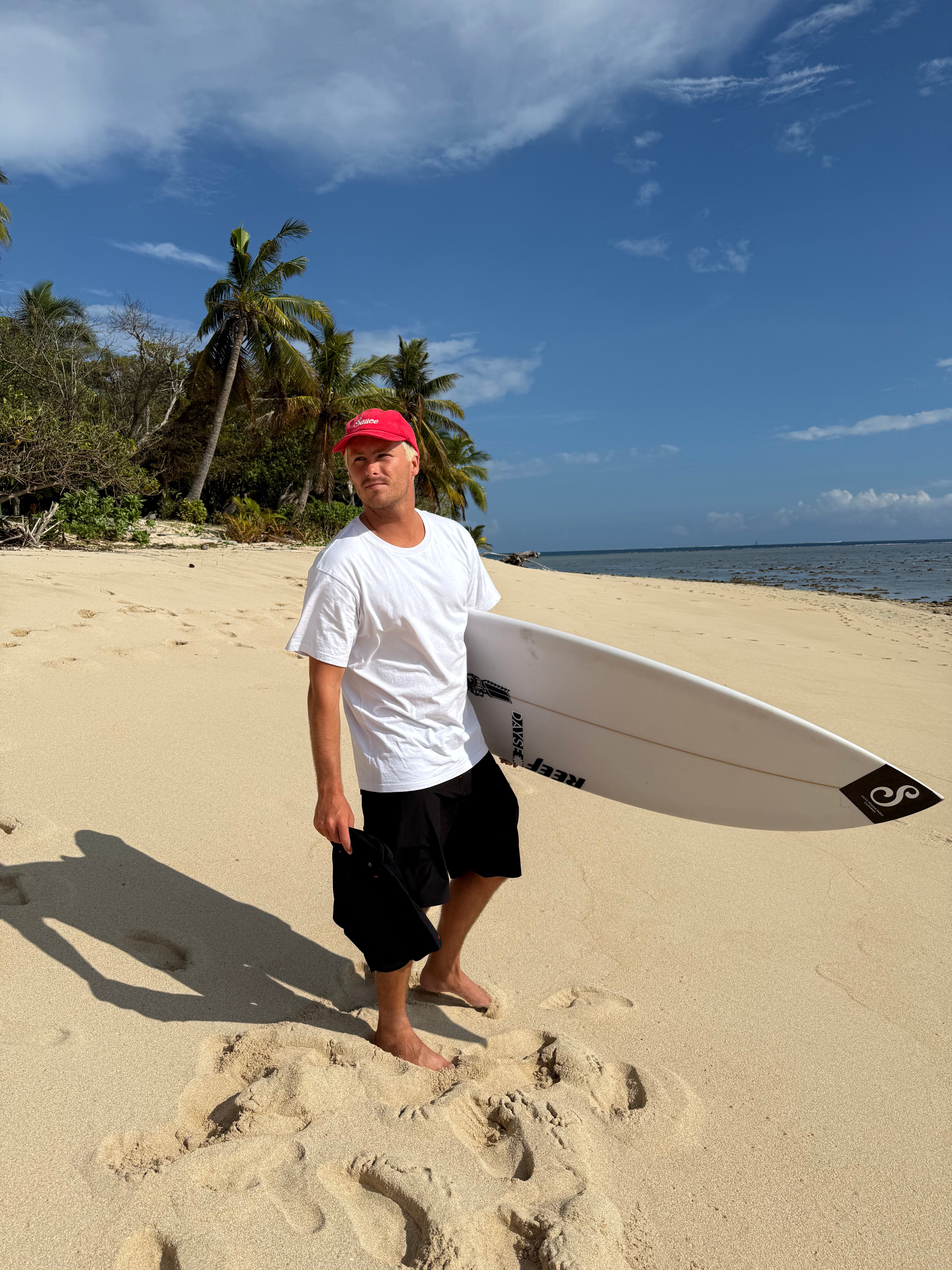 Man holding a surfboard on a sandy beach with palm trees and ocean in the background