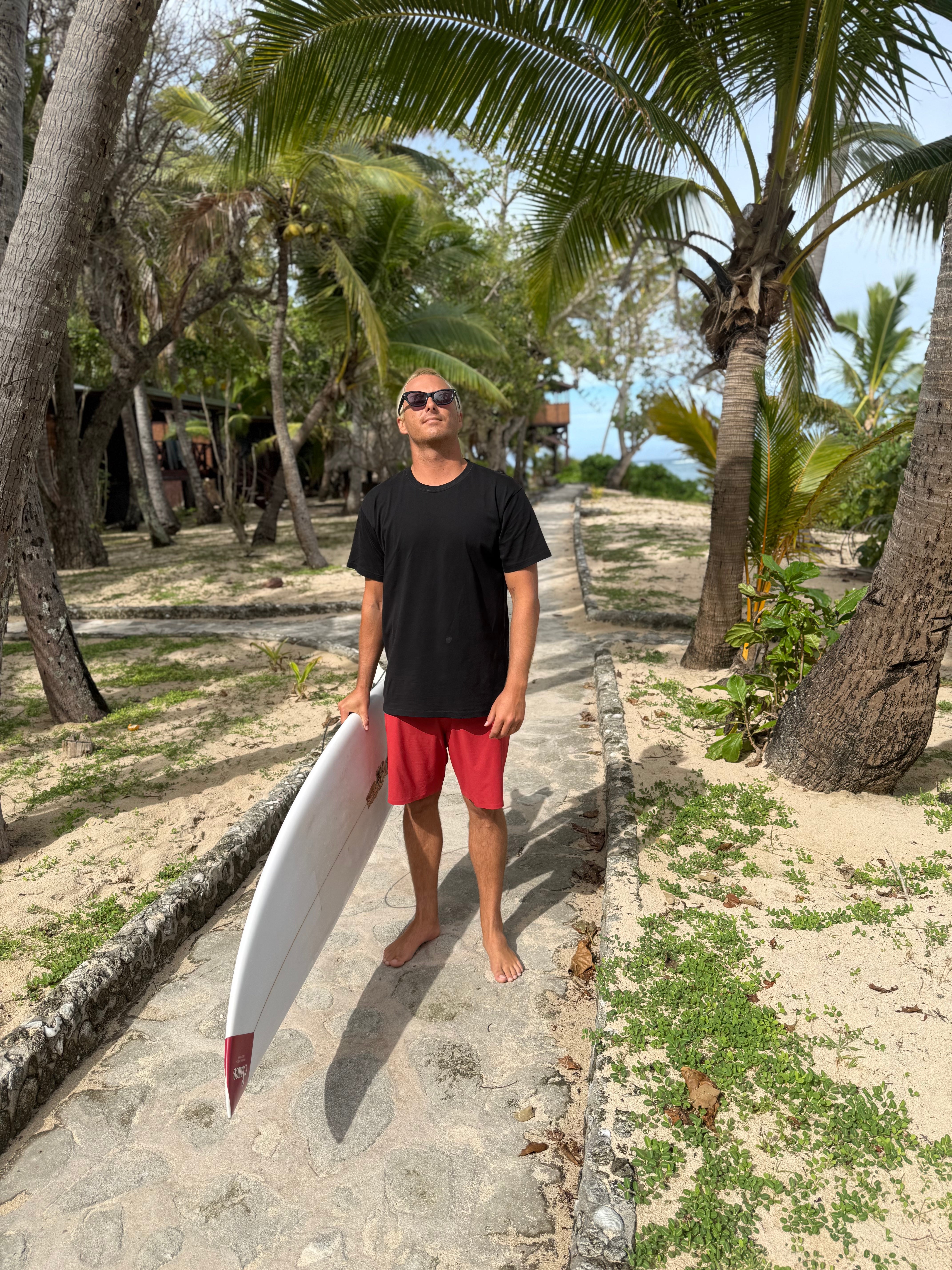 Man holding a surfboard on a sandy path with palm trees in the background