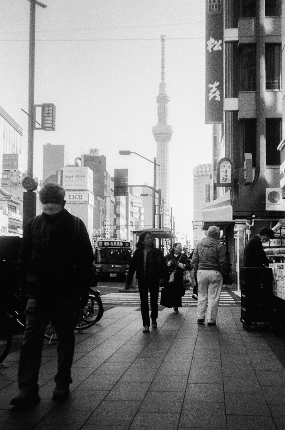 Black and white street scene with people walking on a sidewalk in an urban setting.