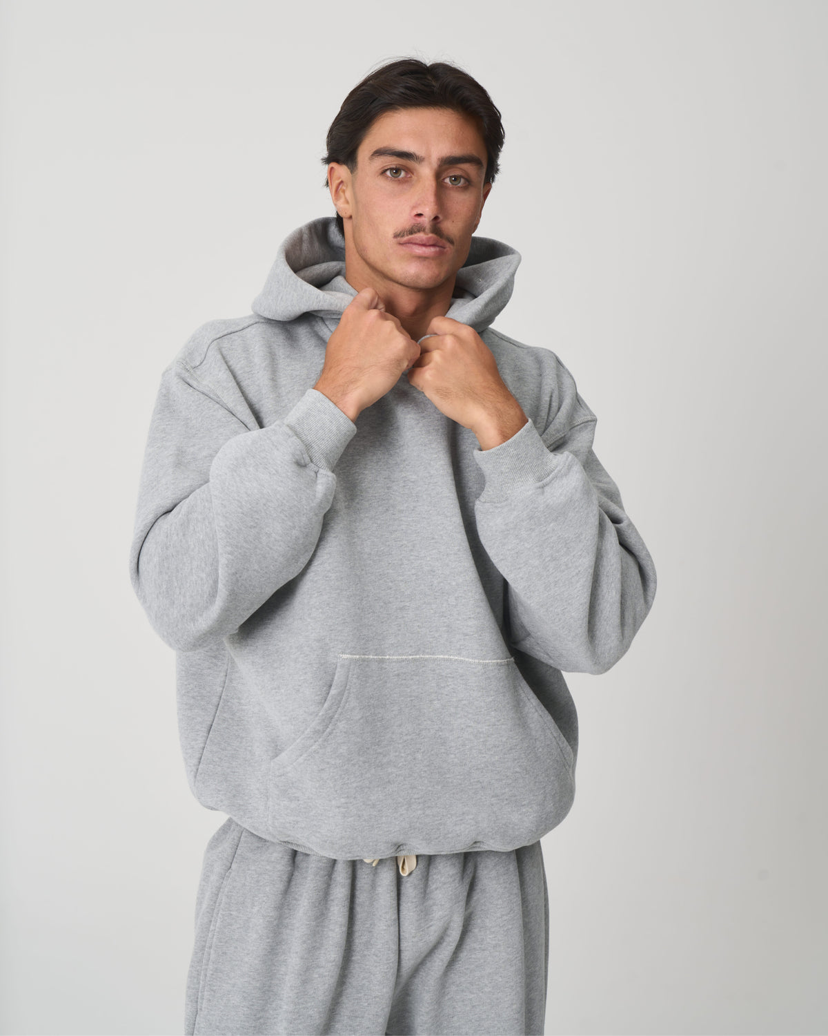 A man wearing a light gray heathered pullover hoodie and matching sweatpants, standing against a light gray studio background.