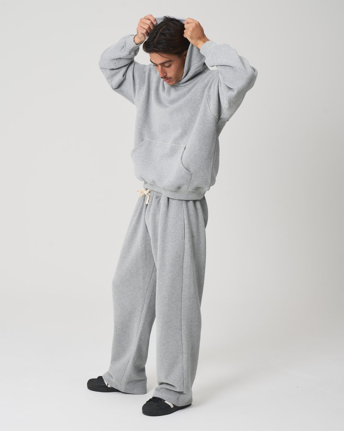 A person modeling a light gray two-piece loungewear set consisting of a pullover hoodie and matching sweatpants, standing against a plain light gray studio background.