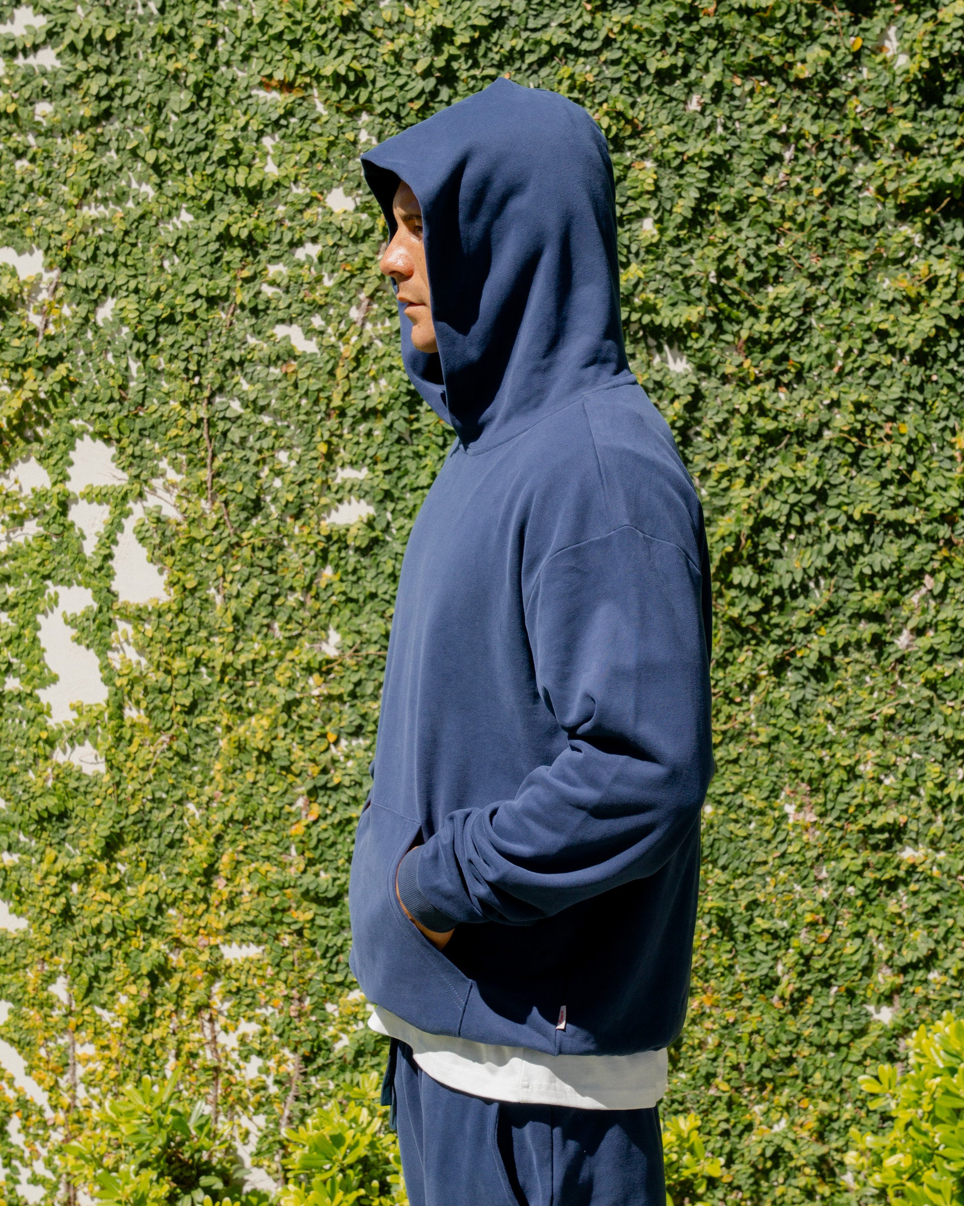 Person wearing a blue hoodie standing against a green hedge.
