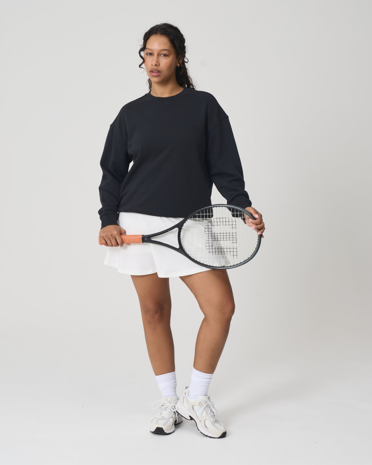 A woman stands against a light gray studio background holding a tennis racket. She is wearing a black long-sleeve top, a white tennis skirt, white socks, and white sneakers.