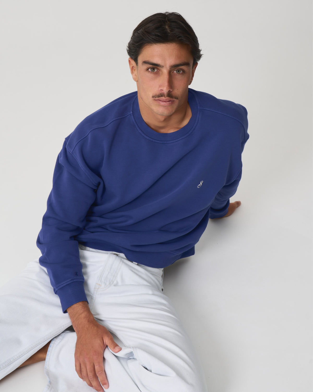 A man wearing a royal blue crewneck sweatshirt and white pants, leaning against a light gray studio background.