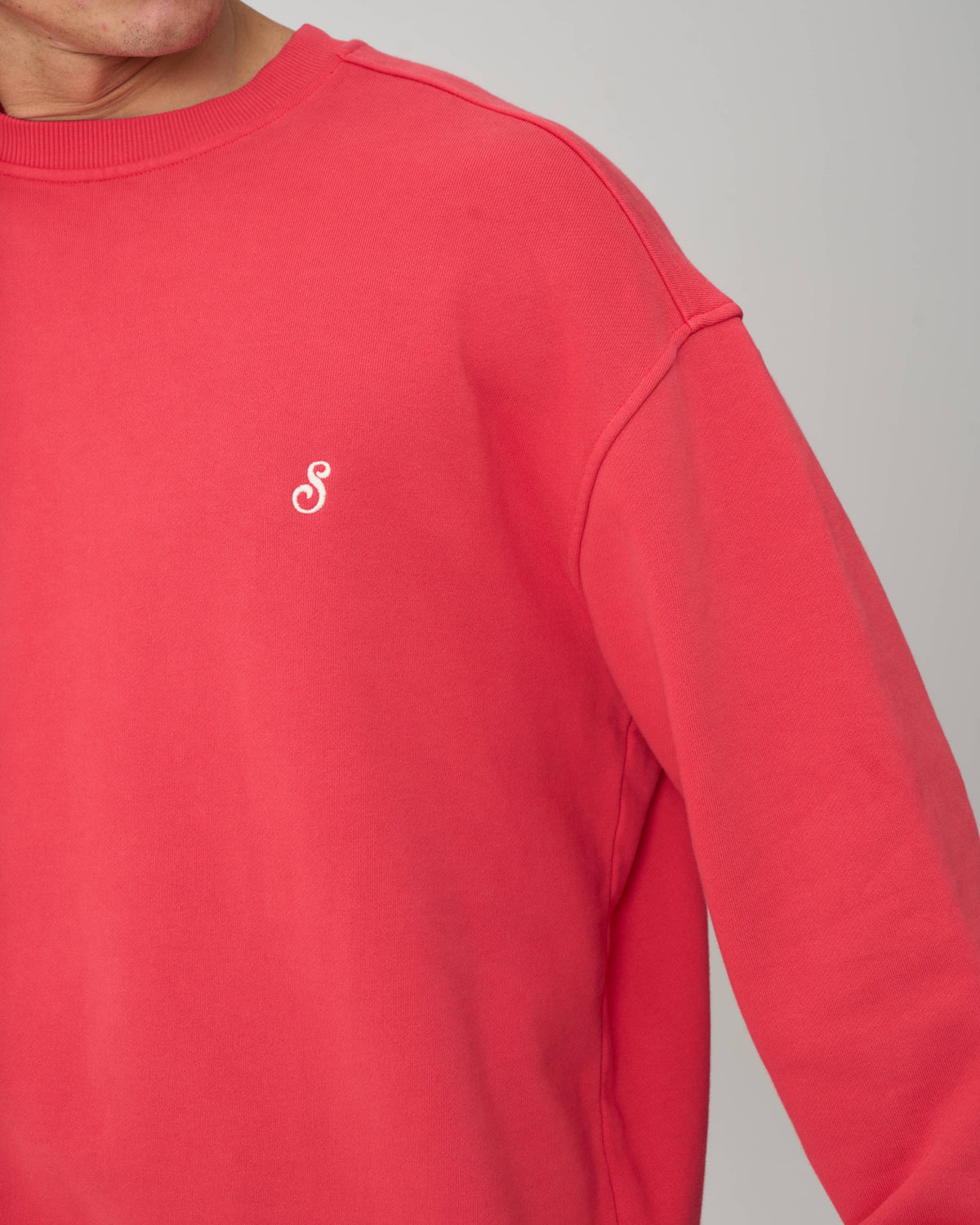 A close-up of a person wearing a coral-red crewneck sweatshirt with a small white embroidered script logo on the left chest, set against a plain light gray background.