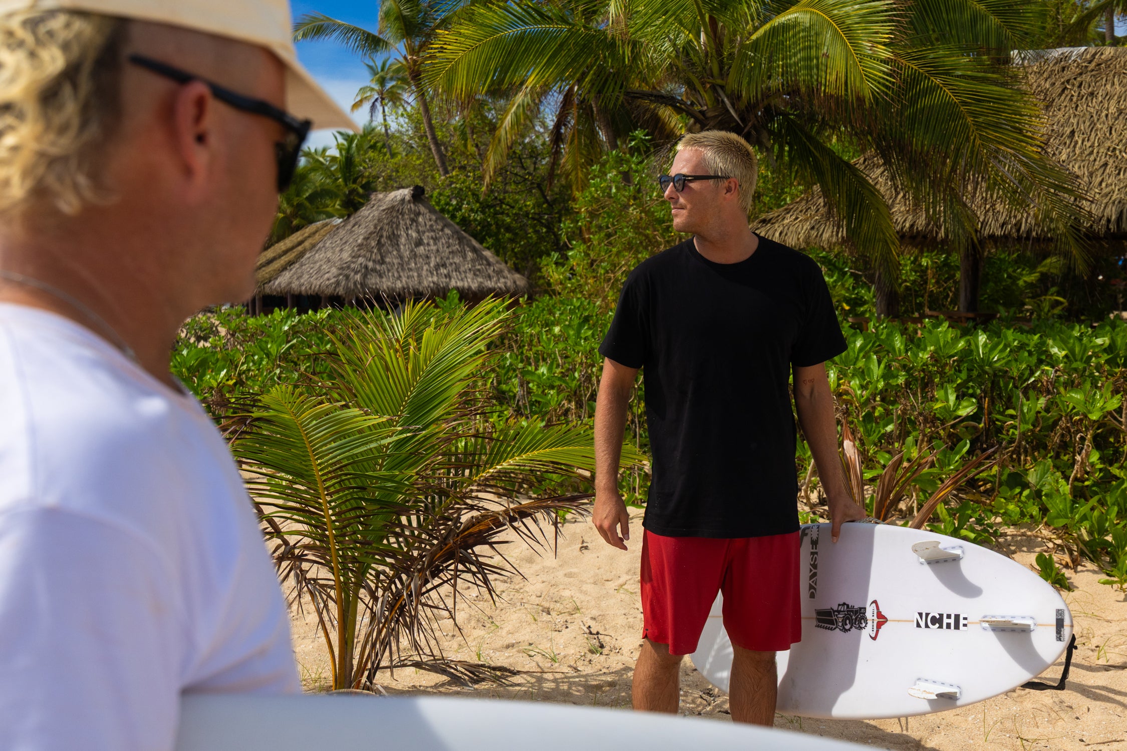 Two men with surfboards on a beach with palm trees and thatched huts in the background.