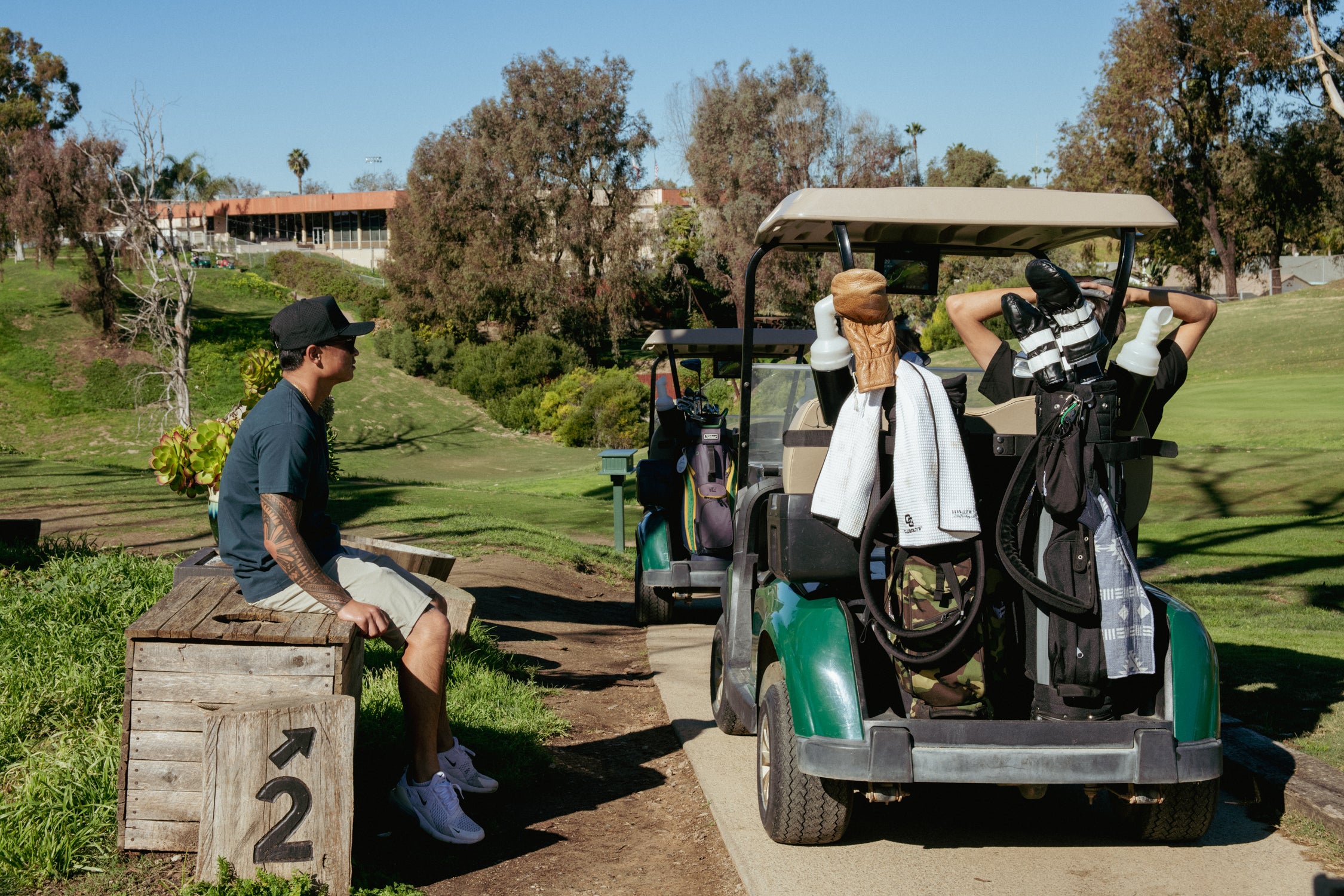 Two men on a golf course with one sitting on a wooden box and the other in a golf cart.