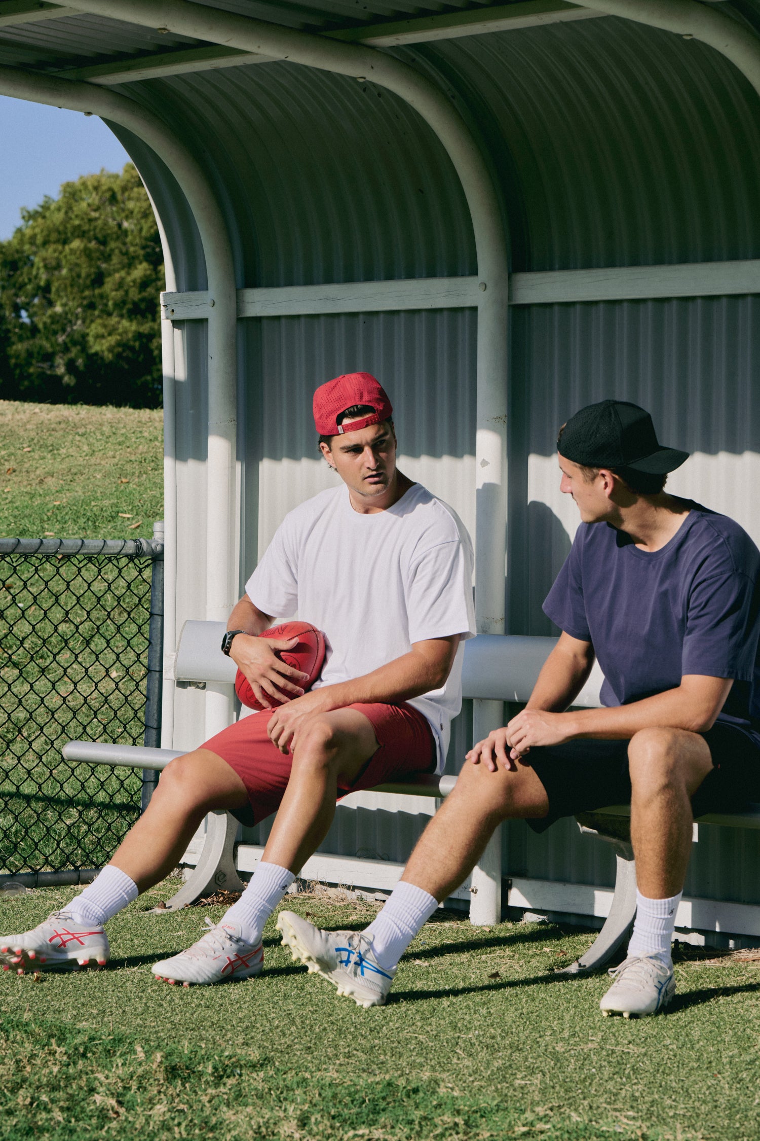 Two young men sitting on a bench outdoors, one holding a sports ball.