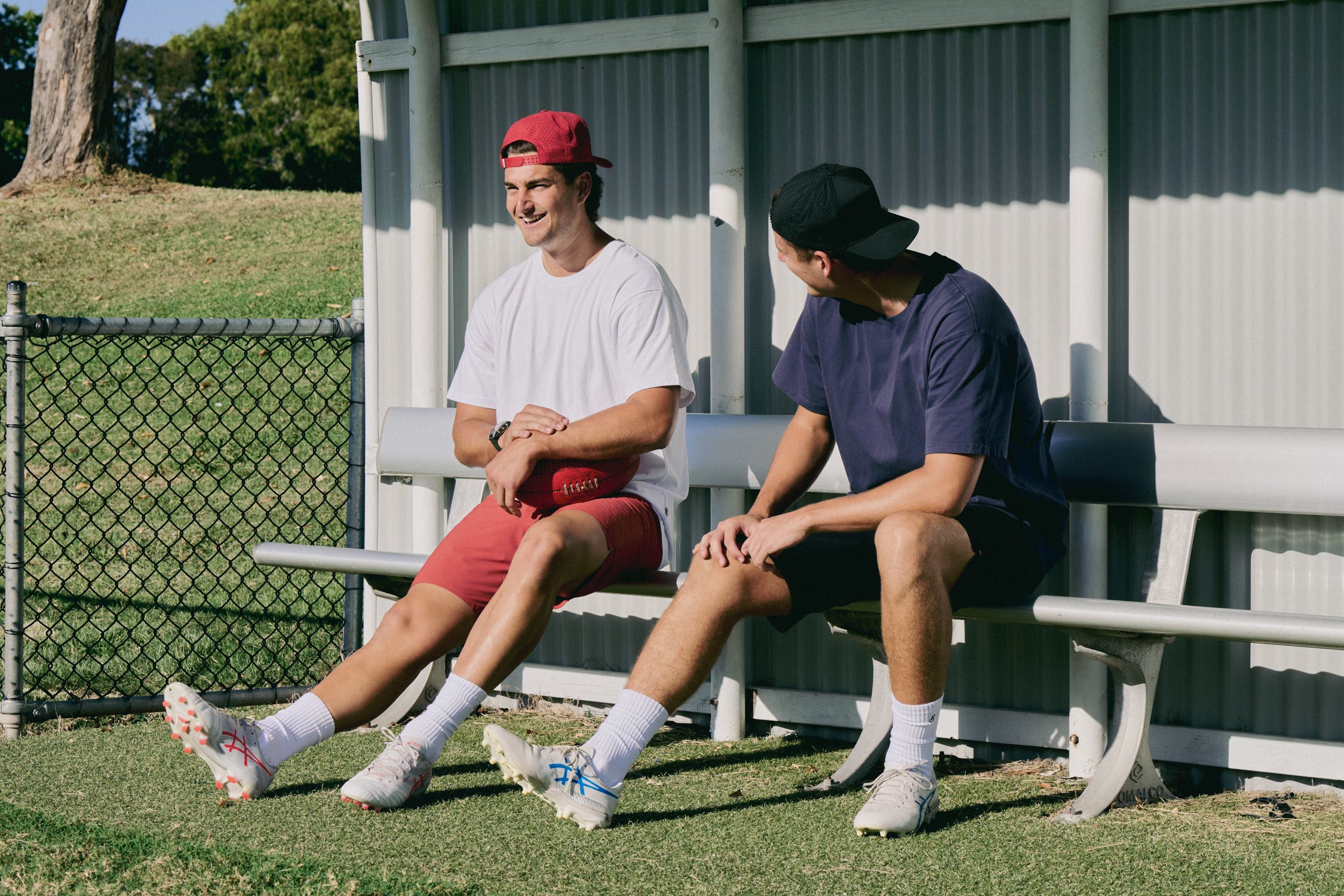 Two young men sitting on a bench outdoors, one wearing a red cap and white shirt, the other in a black cap and dark shirt.
