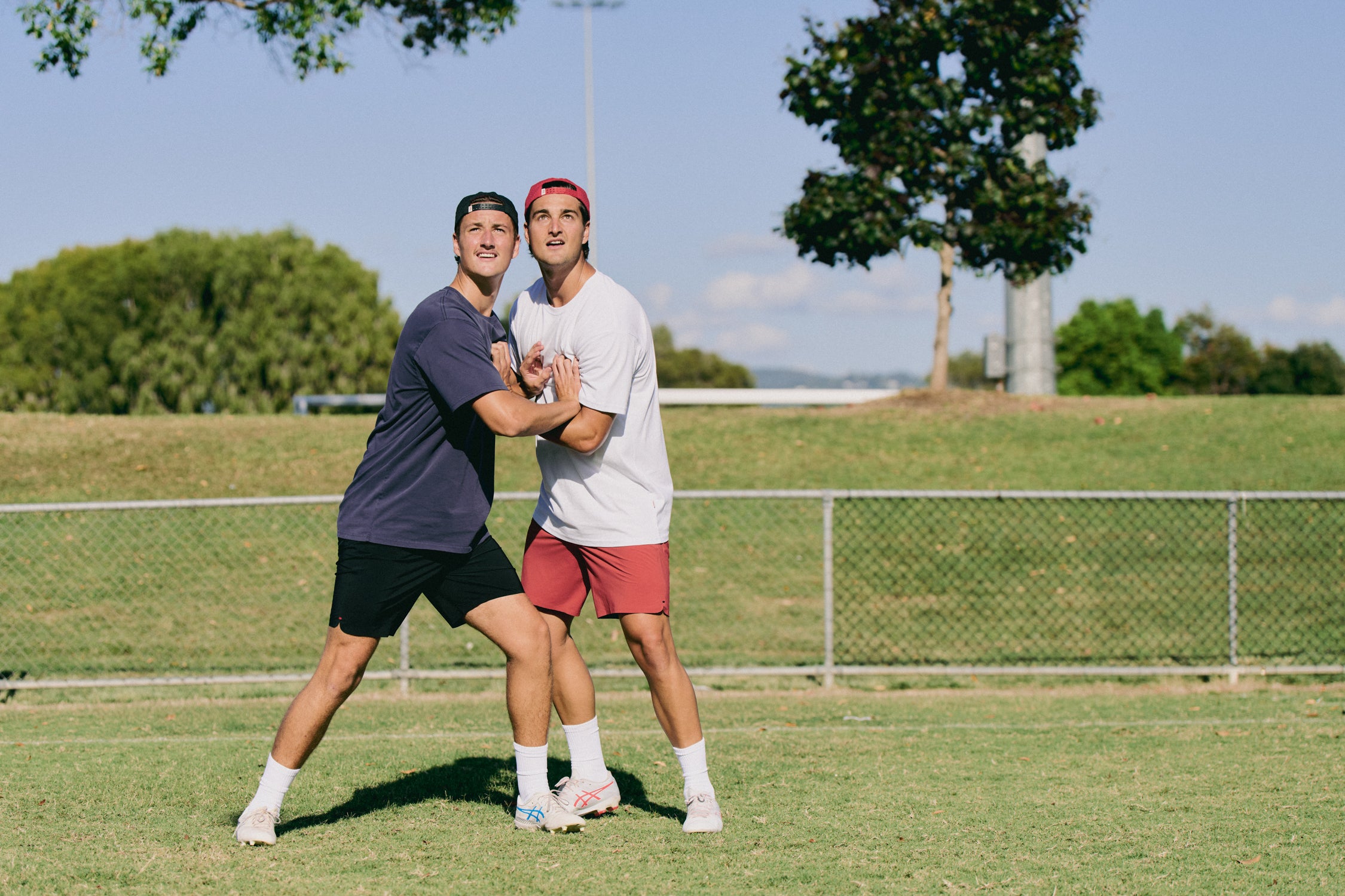 Two young men playing a game of tag on a grassy field with trees and a fence in the background.