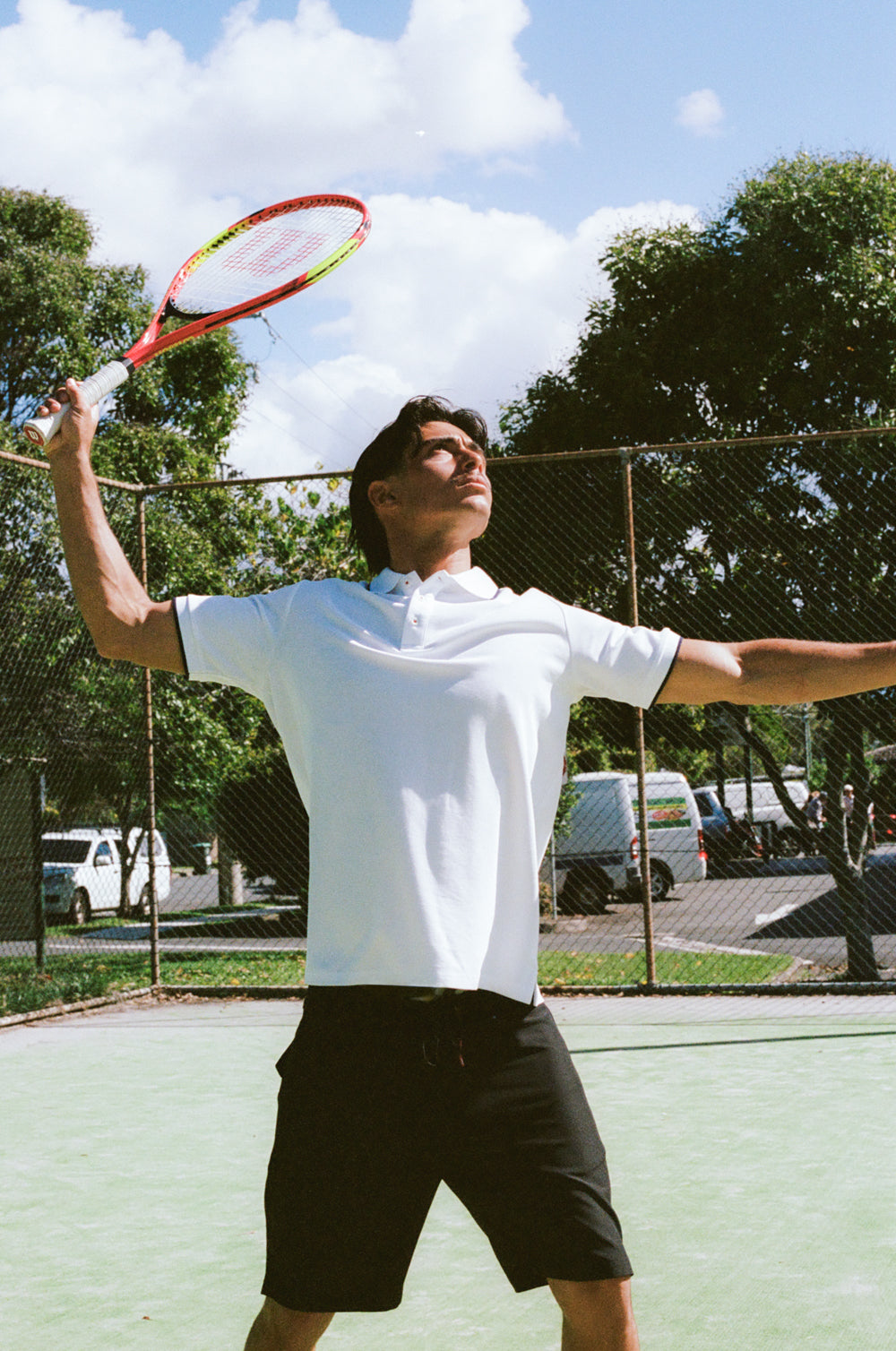 Person playing tennis on a court with trees and cars in the background