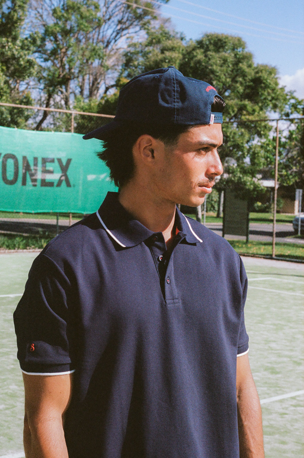 Man wearing a navy blue cap and polo shirt on a tennis court with a green banner in the background.