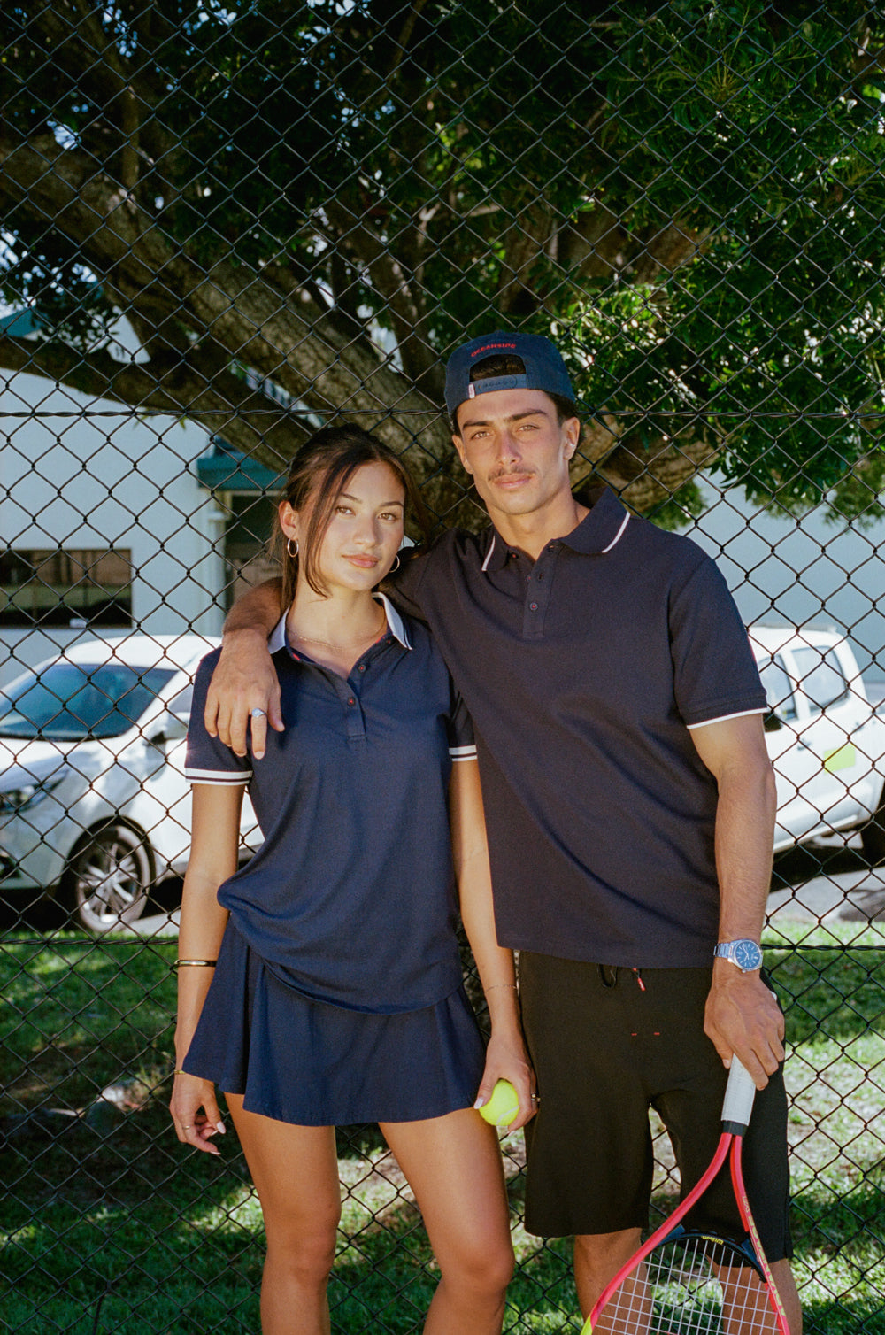 Two people posing together with a tennis racket in front of a chain-link fence.