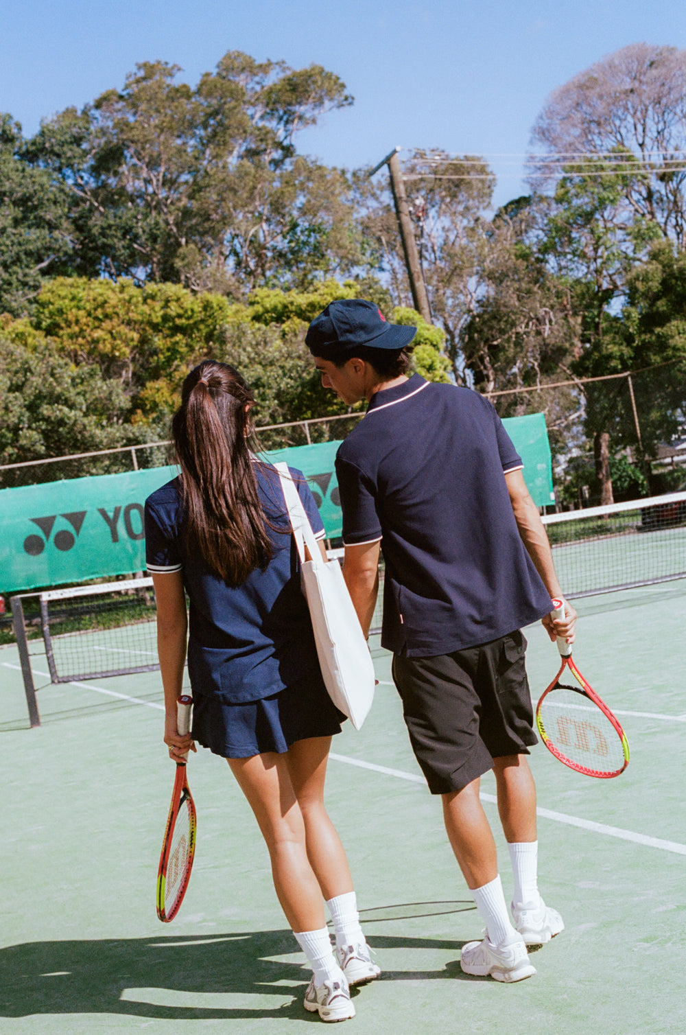 Two people on a tennis court holding tennis rackets with a Yonex logo in the background.