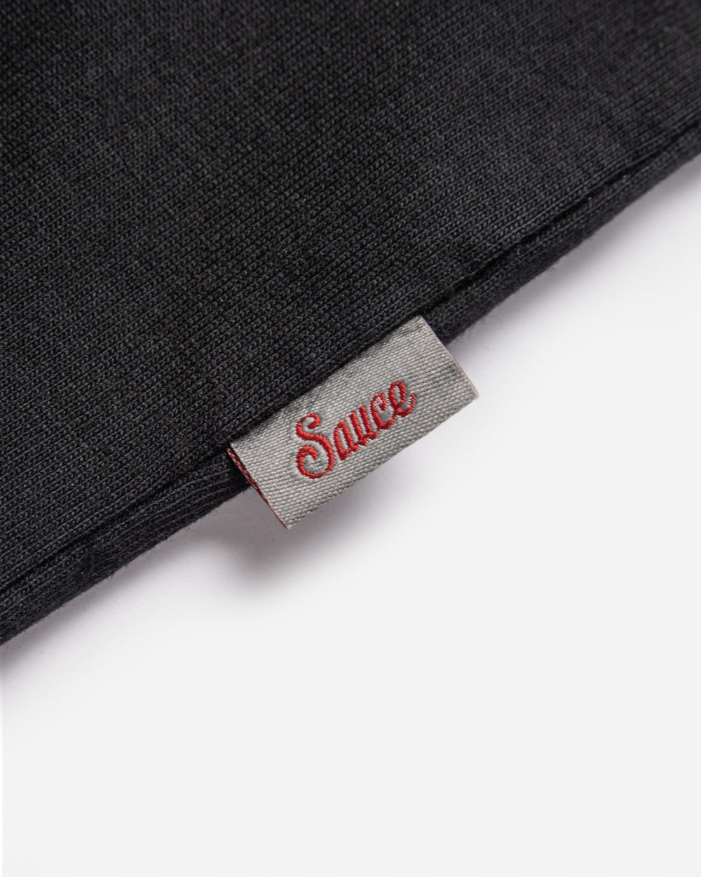 Close-up of a black fabric patch featuring the 'Sauce' label in red stitching on a gray background.