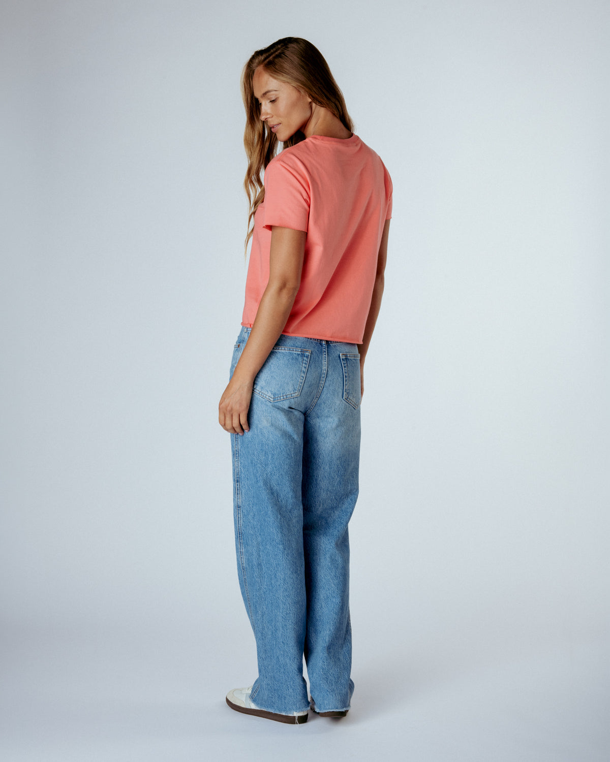 A woman shown from the back wearing a coral pink short-sleeve t-shirt and light-wash blue wide-leg jeans, standing against a light gray studio background.