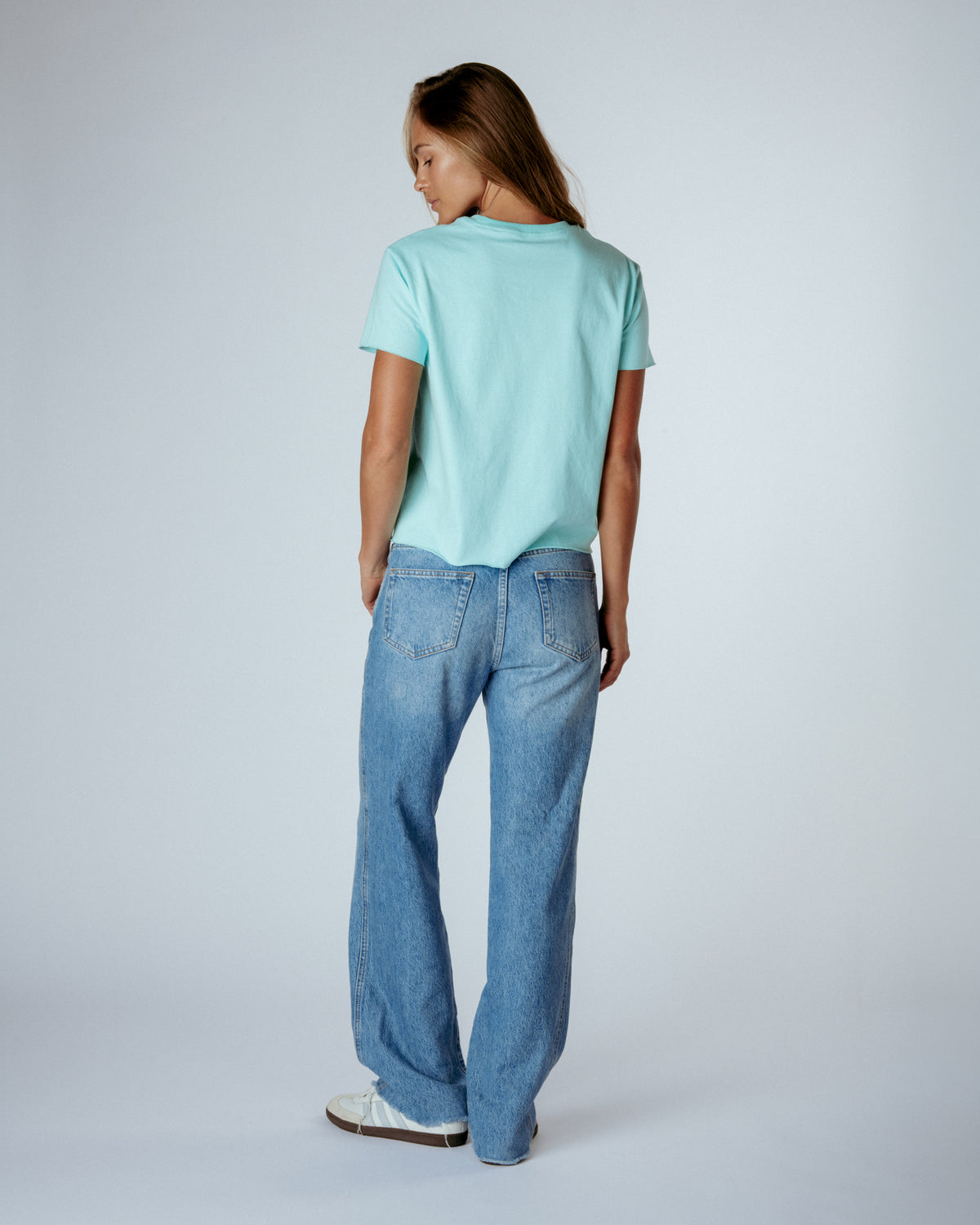 A woman is shown from behind wearing a light mint green t-shirt and light-wash blue jeans, standing against a plain light gray studio background.