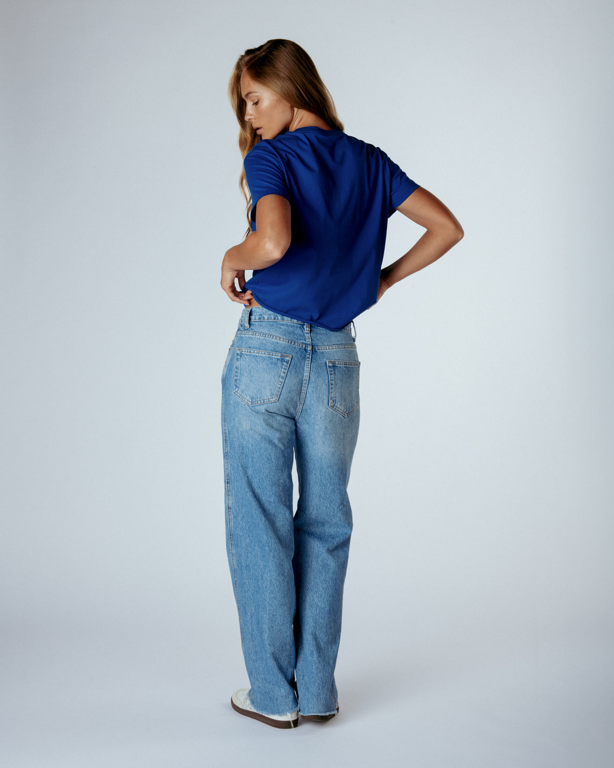 A woman wearing a royal blue short-sleeve t-shirt and light-wash wide-leg jeans, shown from the back in a studio setting with a light gray background.