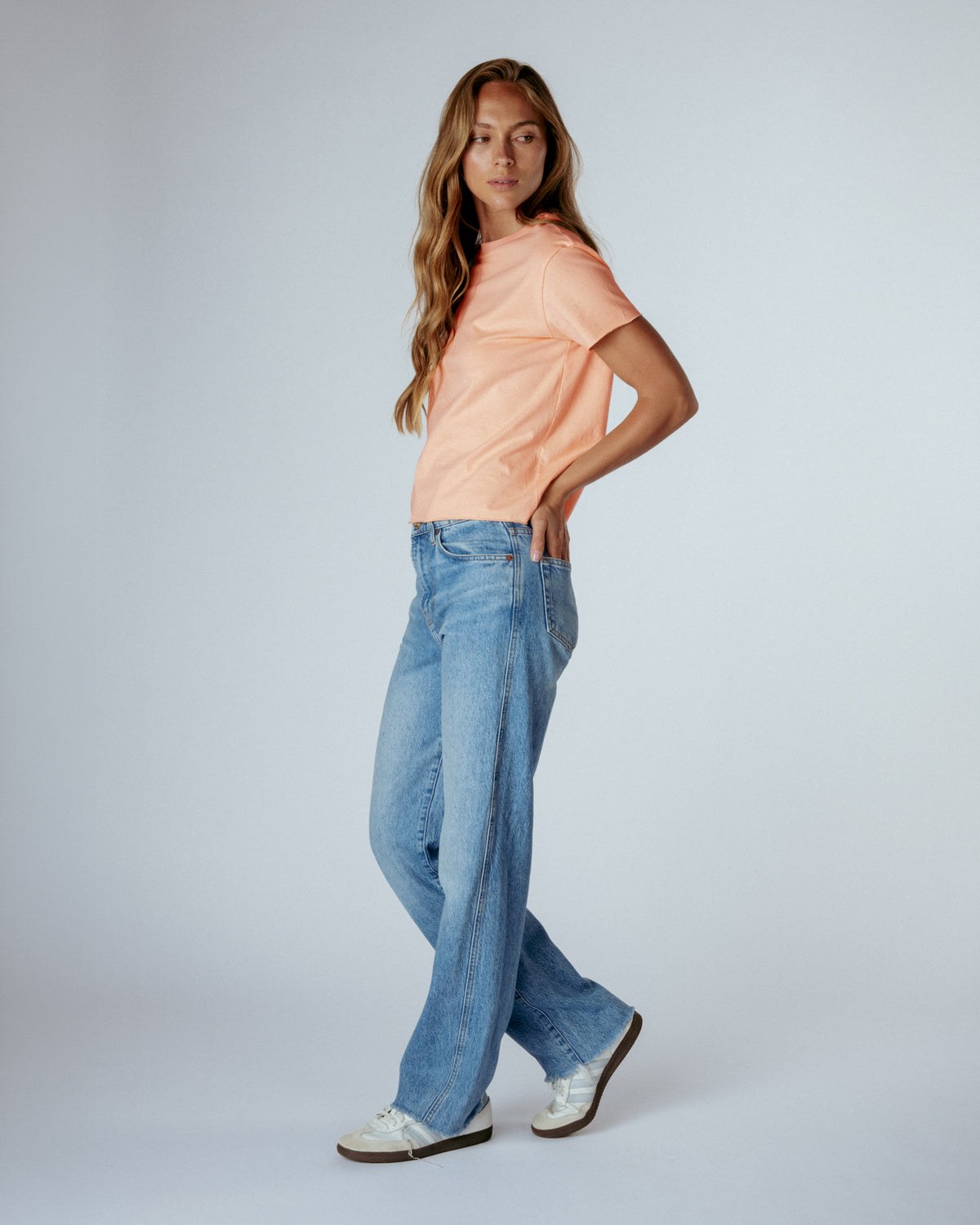 A woman modeling a peach short-sleeve t-shirt and light-wash wide-leg blue jeans, standing against a light gray studio background.