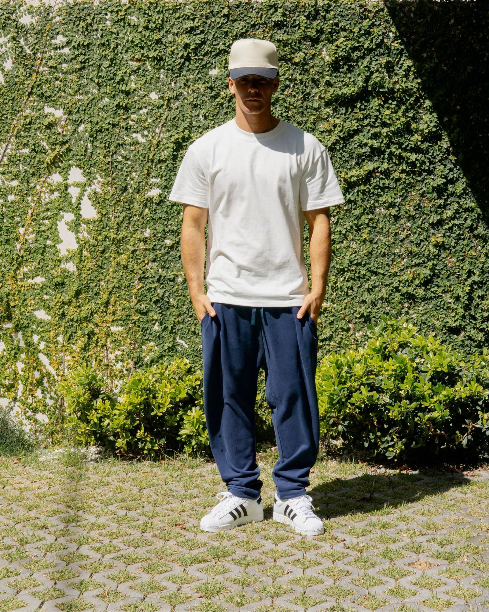 Person wearing a white t-shirt, navy pants, and white sneakers standing in front of greenery.