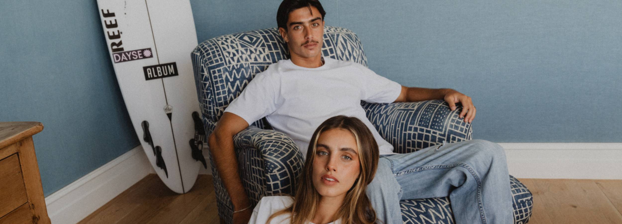 Two people sitting on a patterned chair with a surfboard against a blue wall.