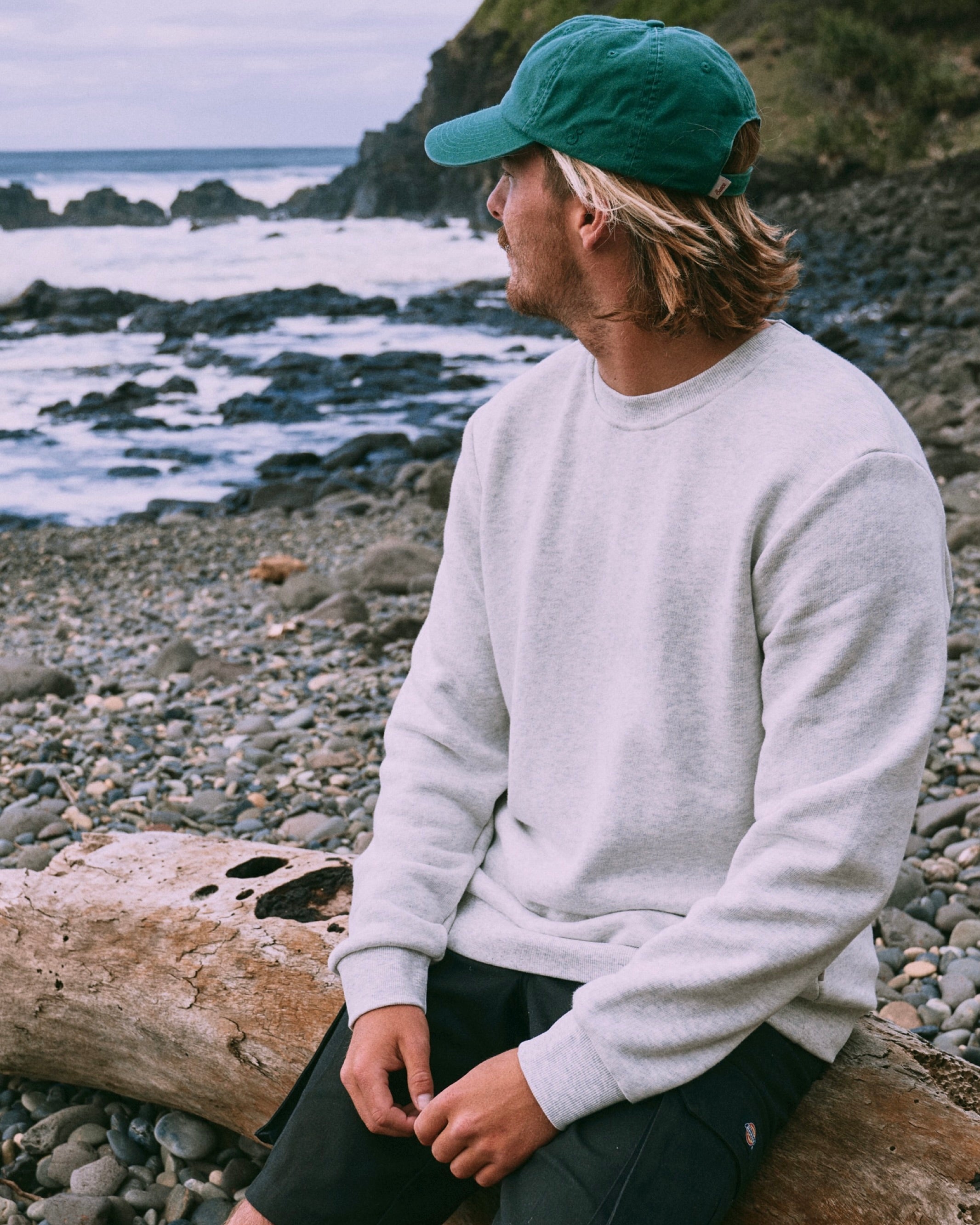 Person sitting on a log by a rocky beach wearing a green cap and light gray sweatshirt.