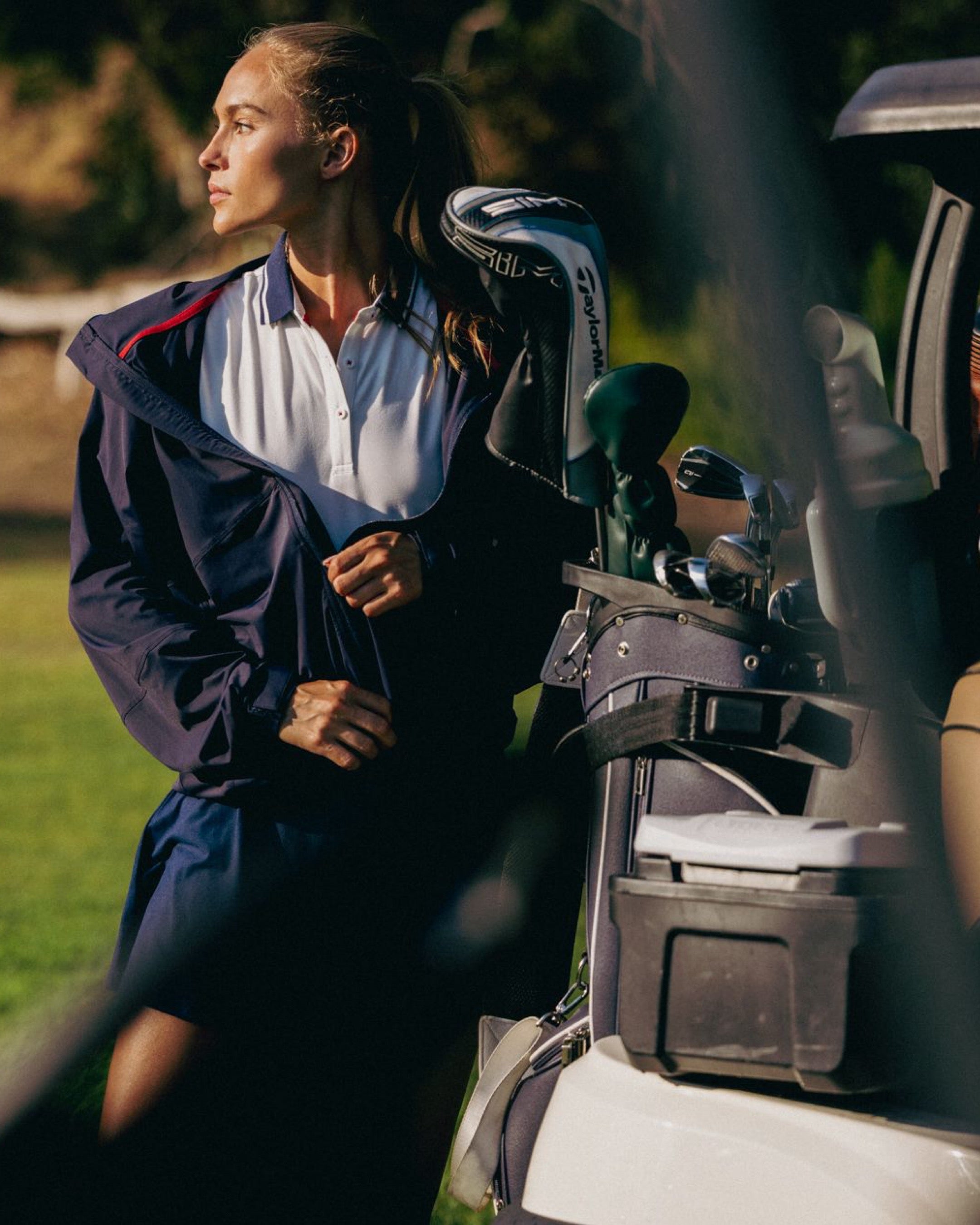 Woman in a navy raincoat standing next to a golf cart with golf clubs on a golf course.