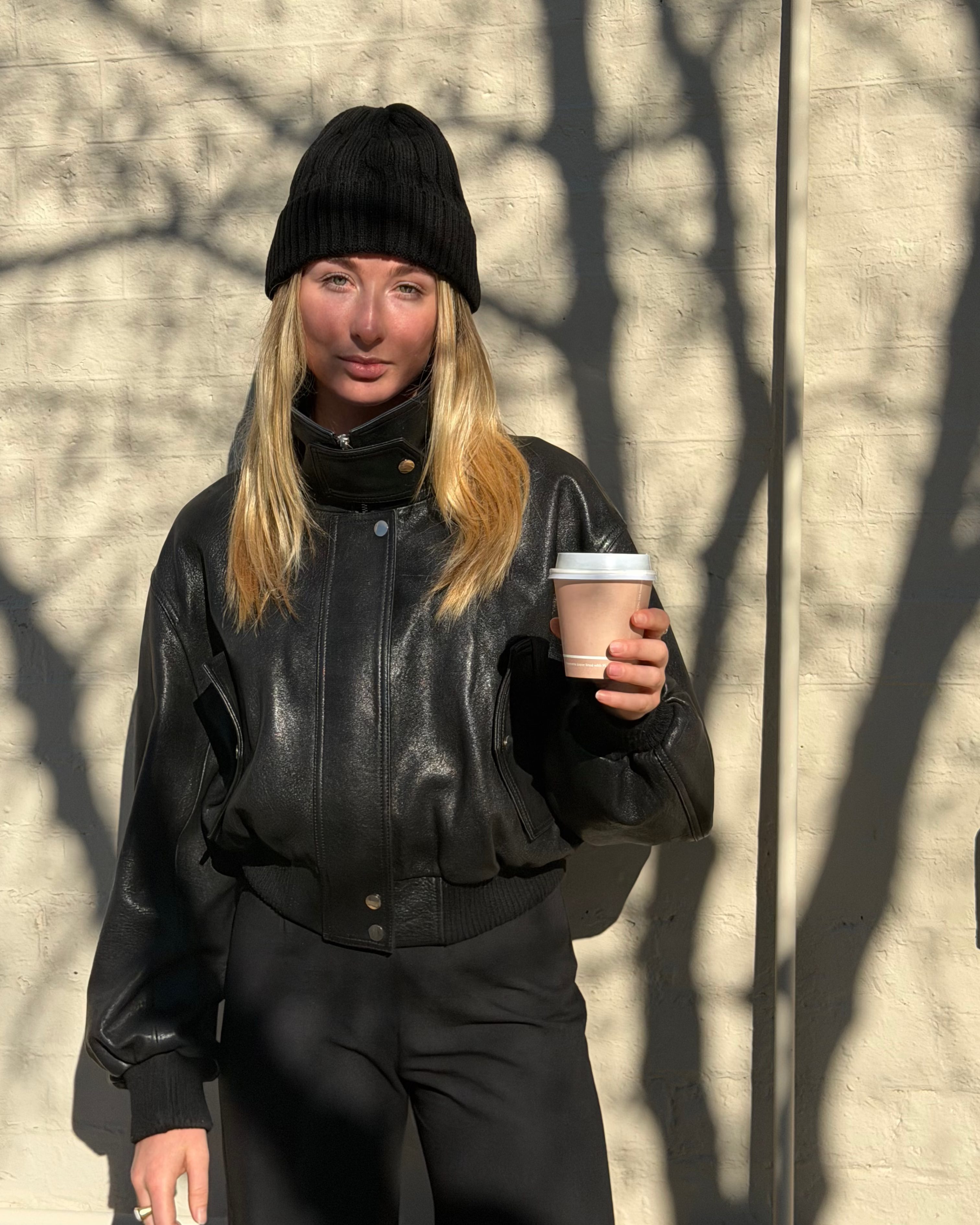 A woman wearing a black leather bomber jacket, black beanie, and black pants holds a takeaway coffee cup against a sunlit beige brick wall with tree shadows.