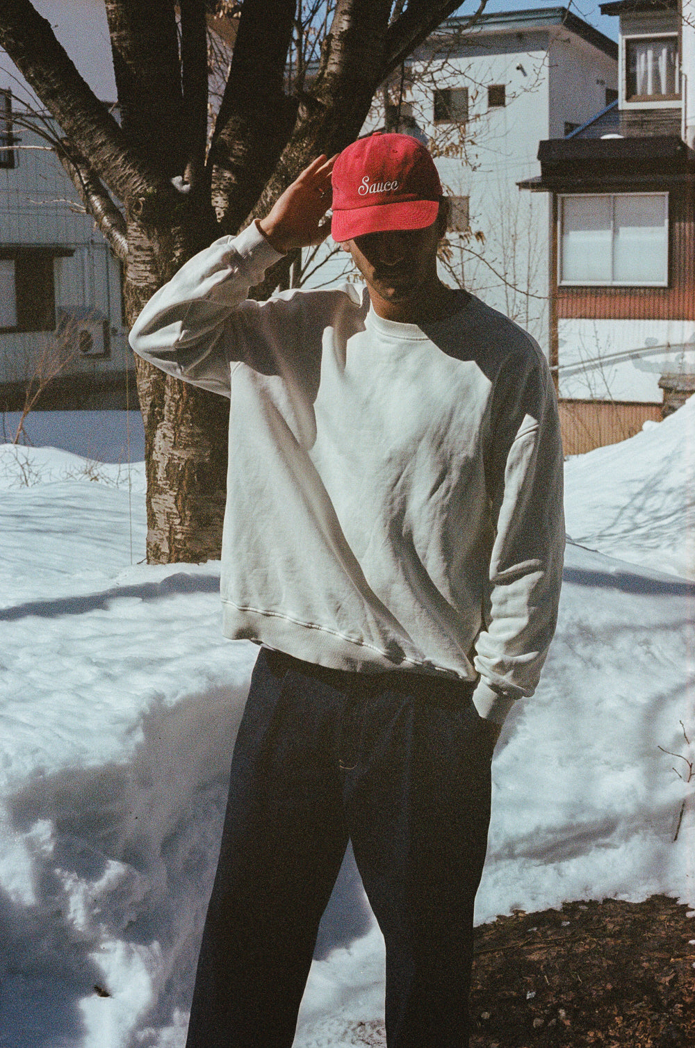 Person wearing a red cap and light gray sweatshirt standing in a snowy landscape with buildings in the background.