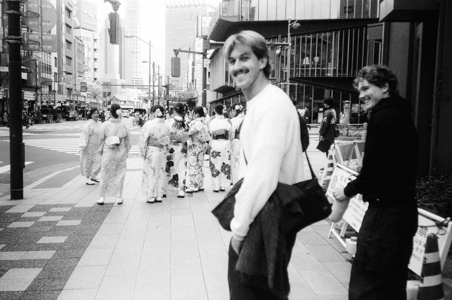 Two men standing on a city street with people in traditional Japanese clothing in the background.