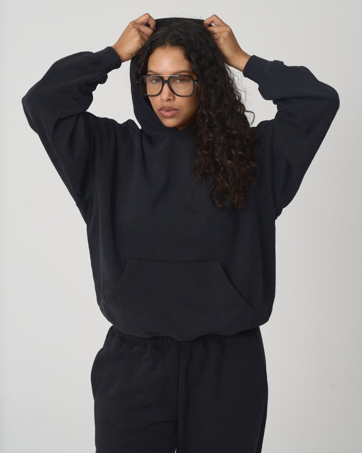 A woman with long curly dark hair and glasses wearing a black oversized hoodie and matching black sweatpants, posing with her hands on her hood against a light gray studio background.