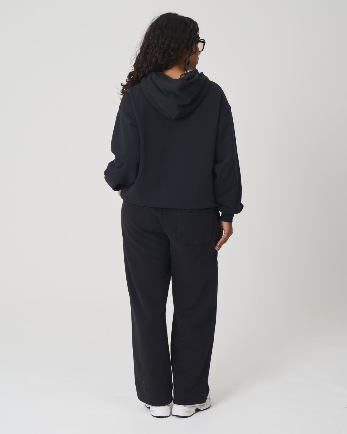 A person shown from the back wearing a black cropped hoodie and matching black wide-leg sweatpants, standing against a light gray studio background.
