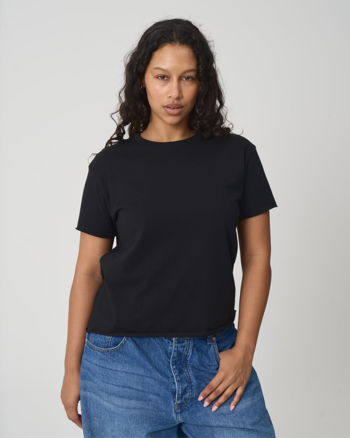 A woman with curly dark hair wearing a plain black short-sleeve crewneck t-shirt and blue jeans, standing against a light gray studio background.