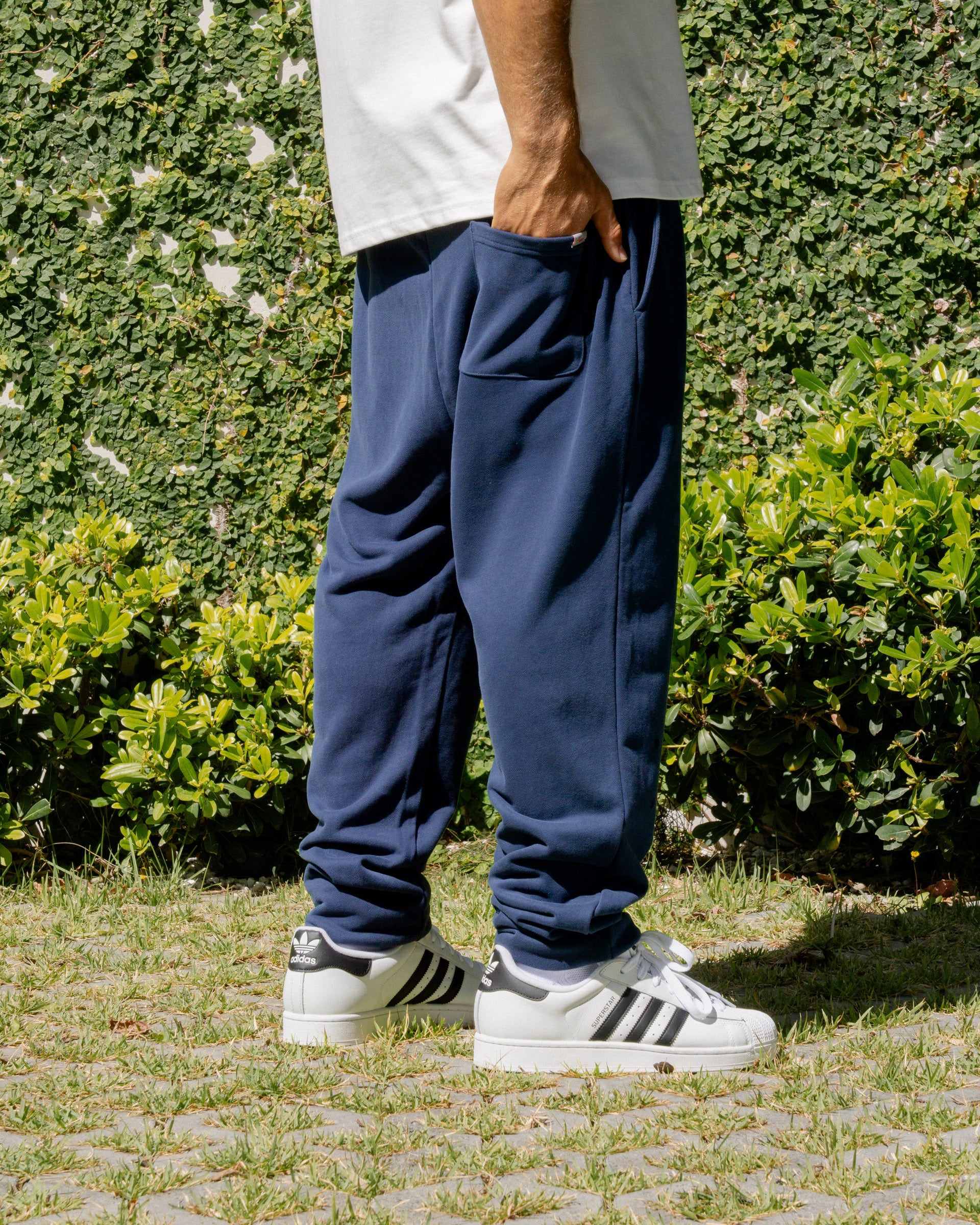 Person wearing navy pants and white Adidas sneakers standing on a grassy area with green bushes in the background.