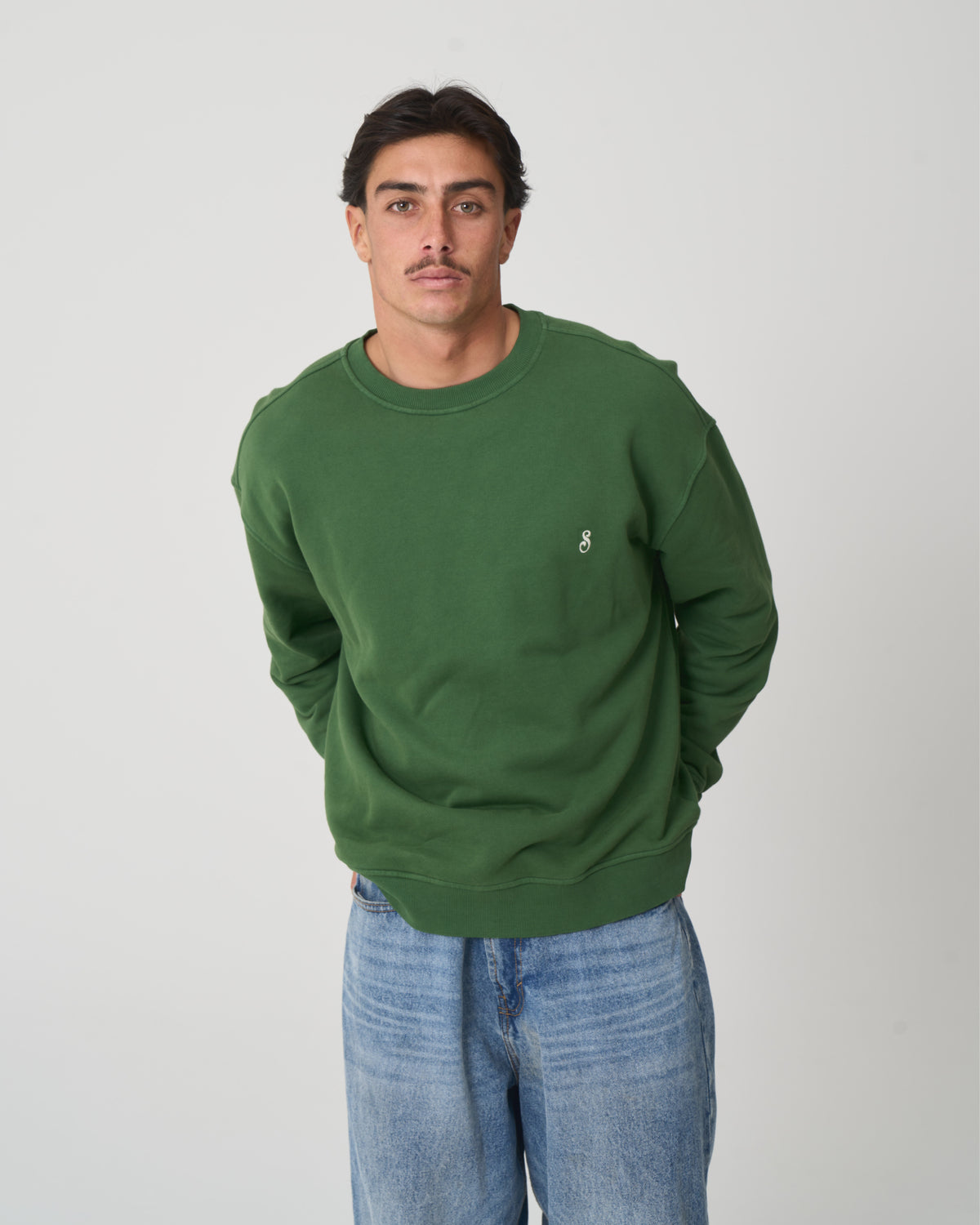 A man wearing a green crewneck sweatshirt with a small white embroidered logo on the left chest, paired with light blue jeans, standing against a light gray studio background.