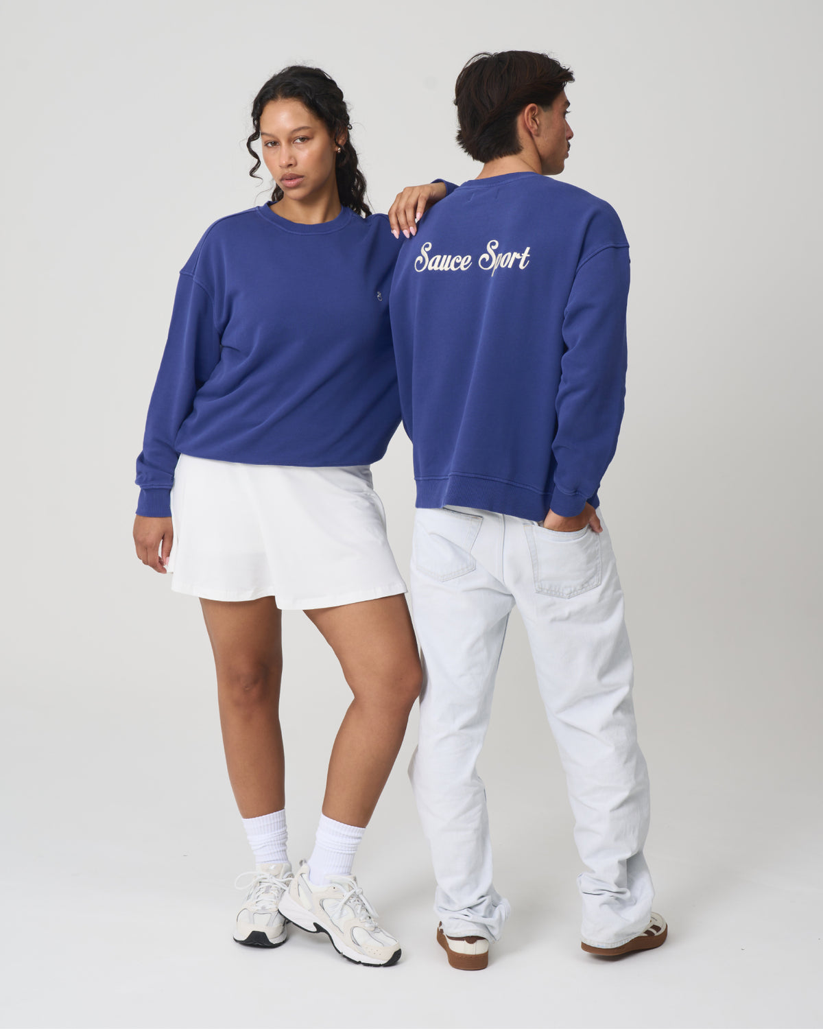Two models wearing a royal blue crewneck sweatshirt with 'Sauce Sport' written in white script on the back, styled with white shorts and white pants against a light gray studio background.