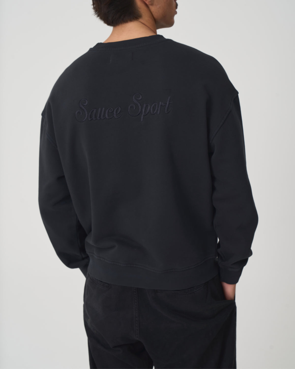 A person shown from the back wearing a black crewneck sweatshirt with 'Sauce Sport' embroidered across the upper back, paired with black pants, against a light gray studio background.