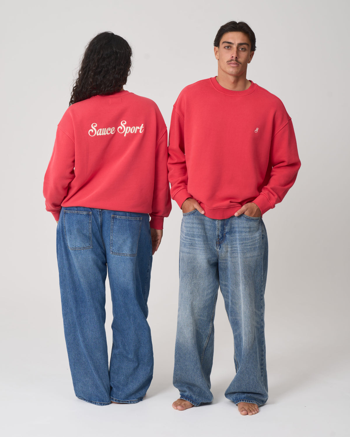 A man and a woman modeling matching coral-red crewneck sweatshirts and loose blue jeans against a plain light gray studio background. The woman shows the back of the sweatshirt with 'Sauce Sport' in white script, and the man shows the front with a small white chest logo.