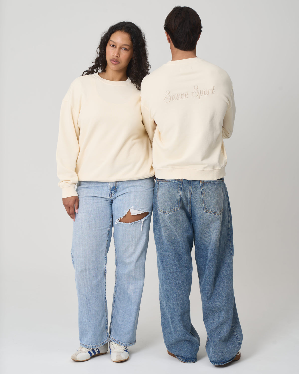 Two models wearing cream-colored crewneck sweatshirts and wide-leg jeans, standing against a light gray studio background. One model is shown from the front and the other from the back, displaying a script logo on the back of the sweatshirt.