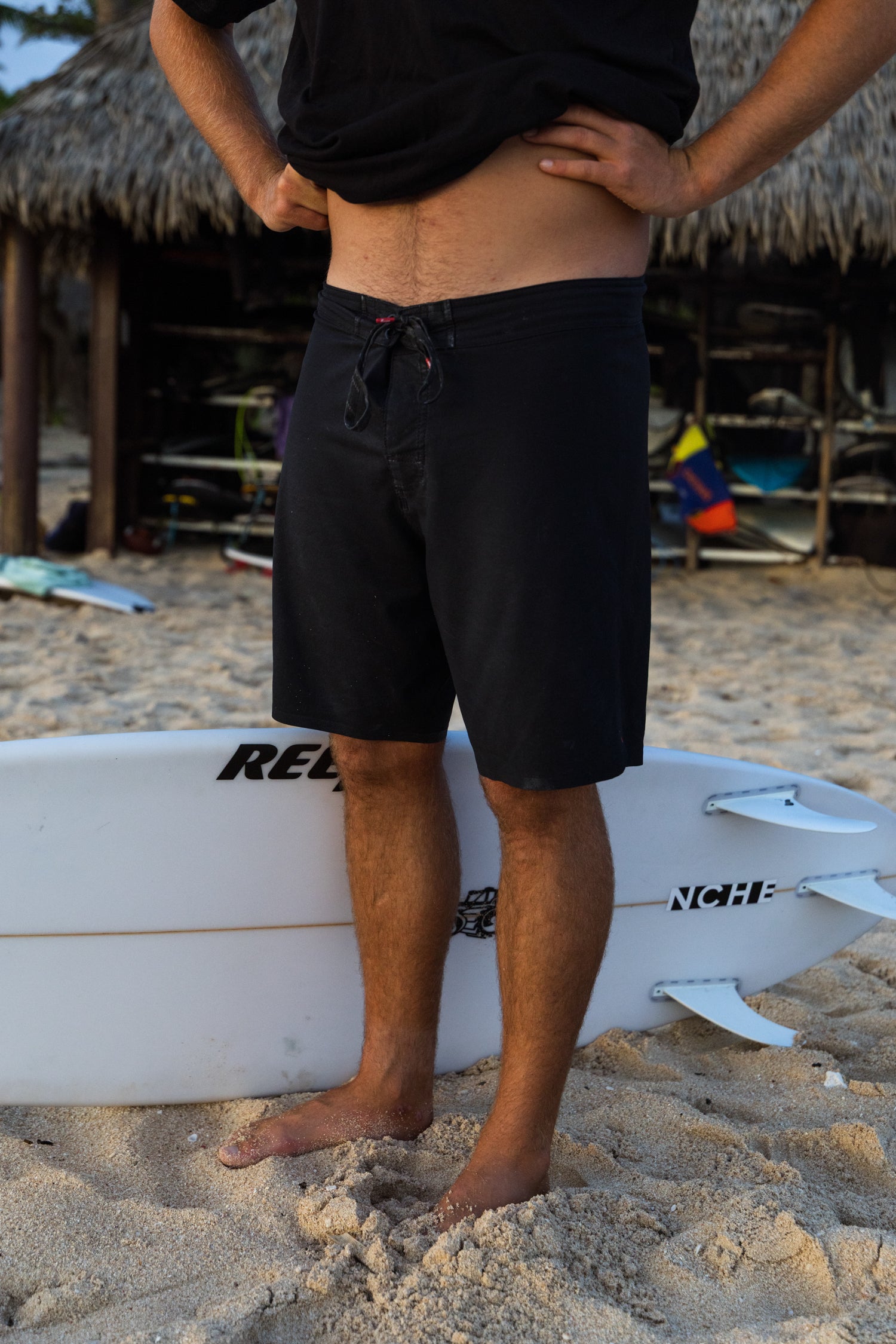 Person wearing black shorts standing on a beach with a white surfboard.