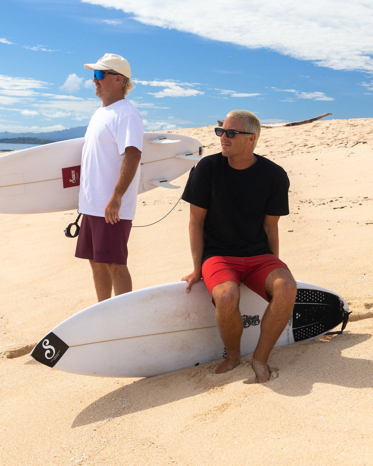 Two men with surfboards on a sandy beach under a blue sky.