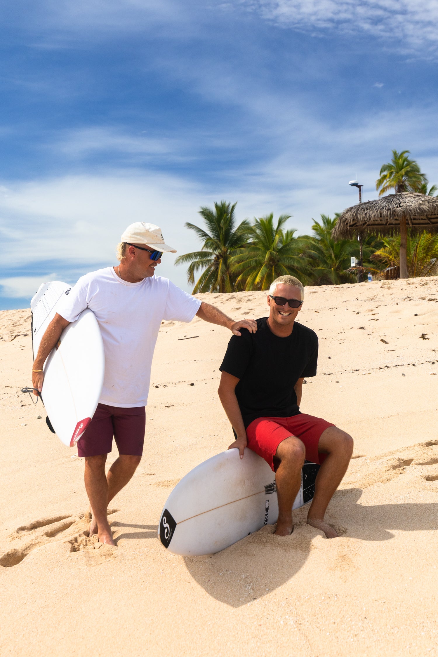 Two men with surfboards on a sandy beach with palm trees and a blue sky.