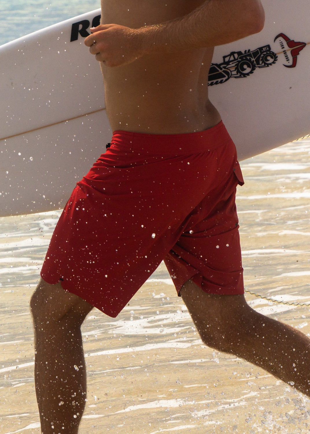 Person wearing red swim shorts holding a white surfboard with branding on a beach.