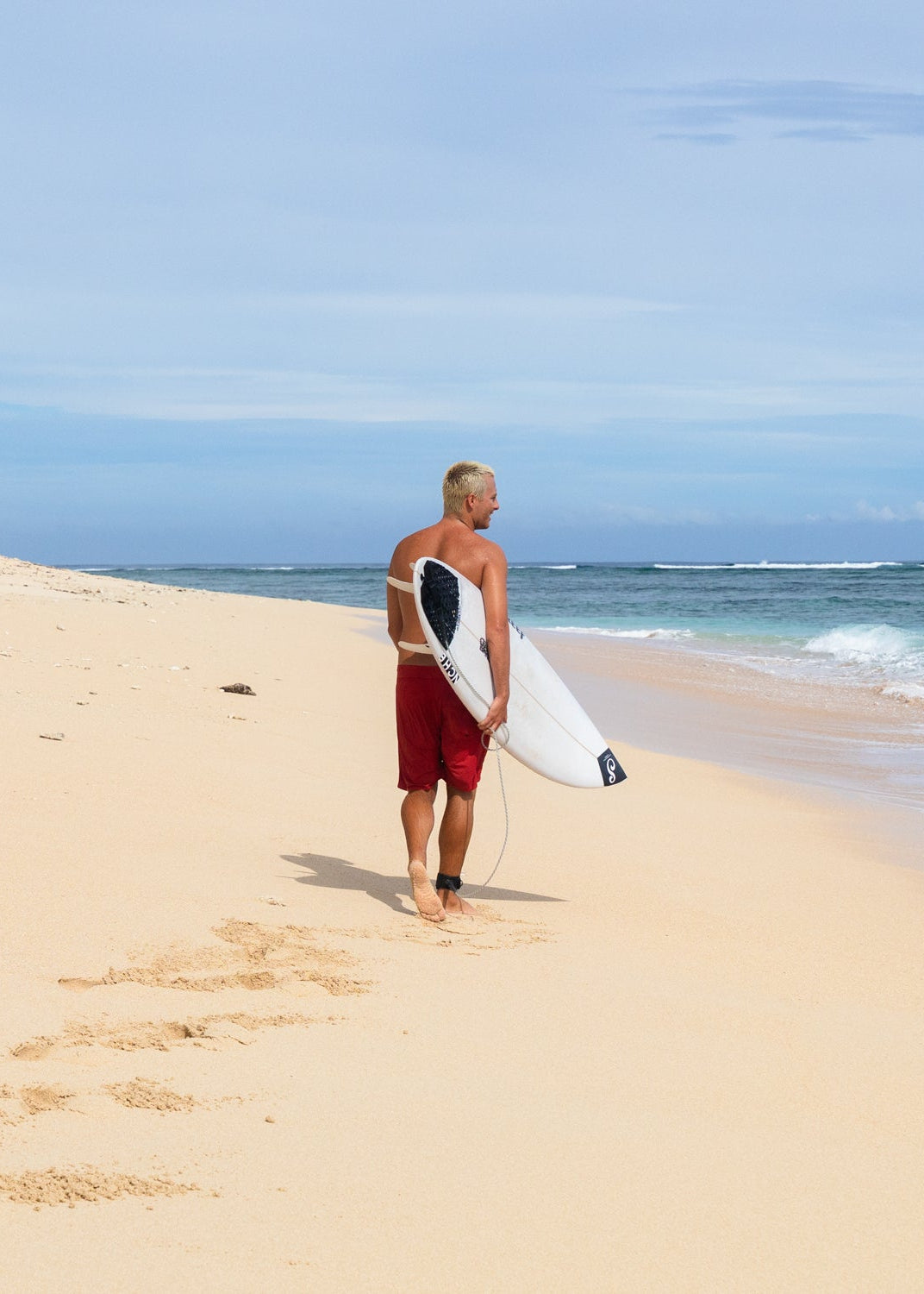 Man holding a surfboard on a sandy beach with ocean and sky in the background