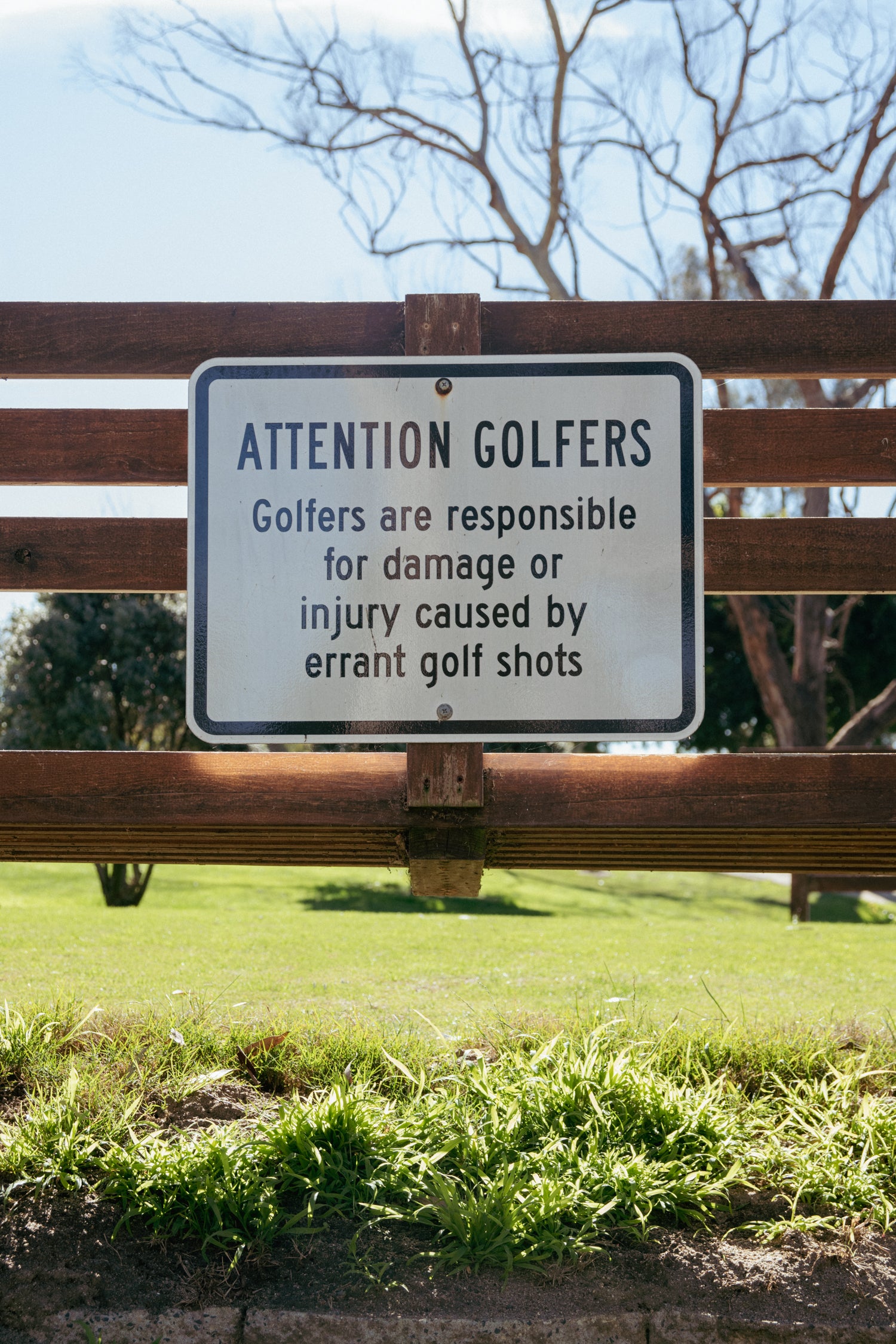 Sign warning golfers about responsibility for damage or injury caused by errant golf shots, attached to a wooden fence.