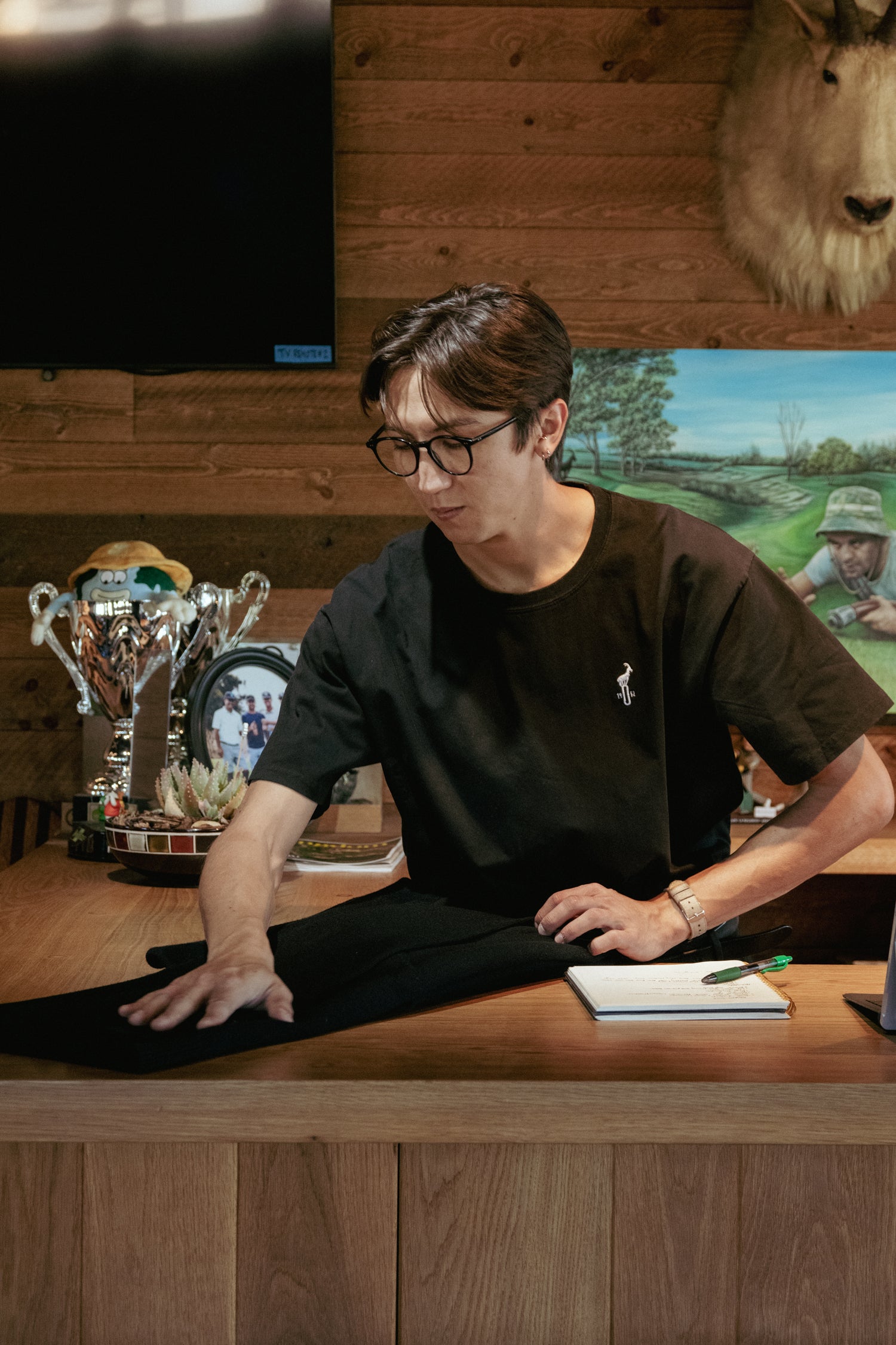 Person in a black shirt sitting at a desk with trophies and a painting in the background