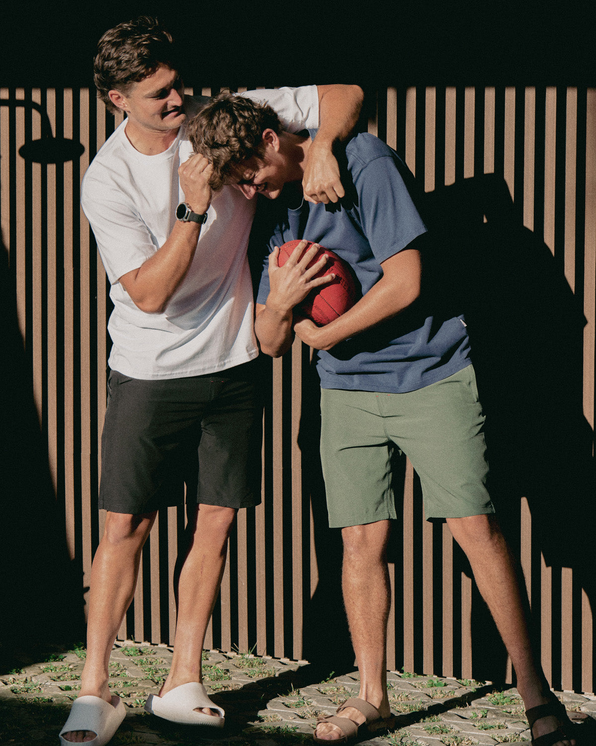 Two young men playing with a red ball outdoors against a striped wall.