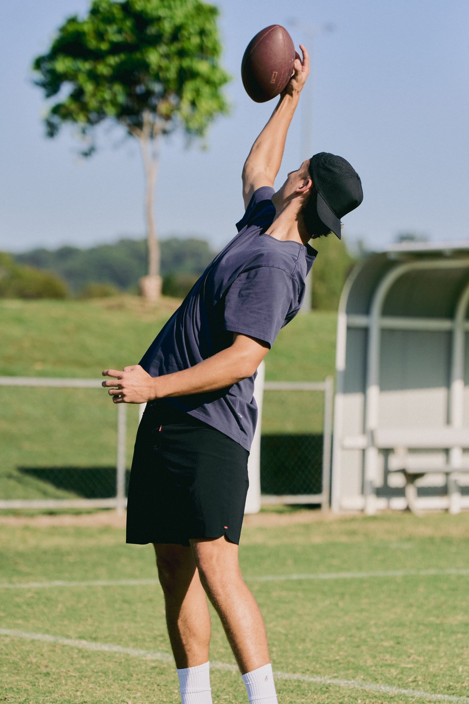 Person catching a football on a sports field with trees and bleachers in the background
